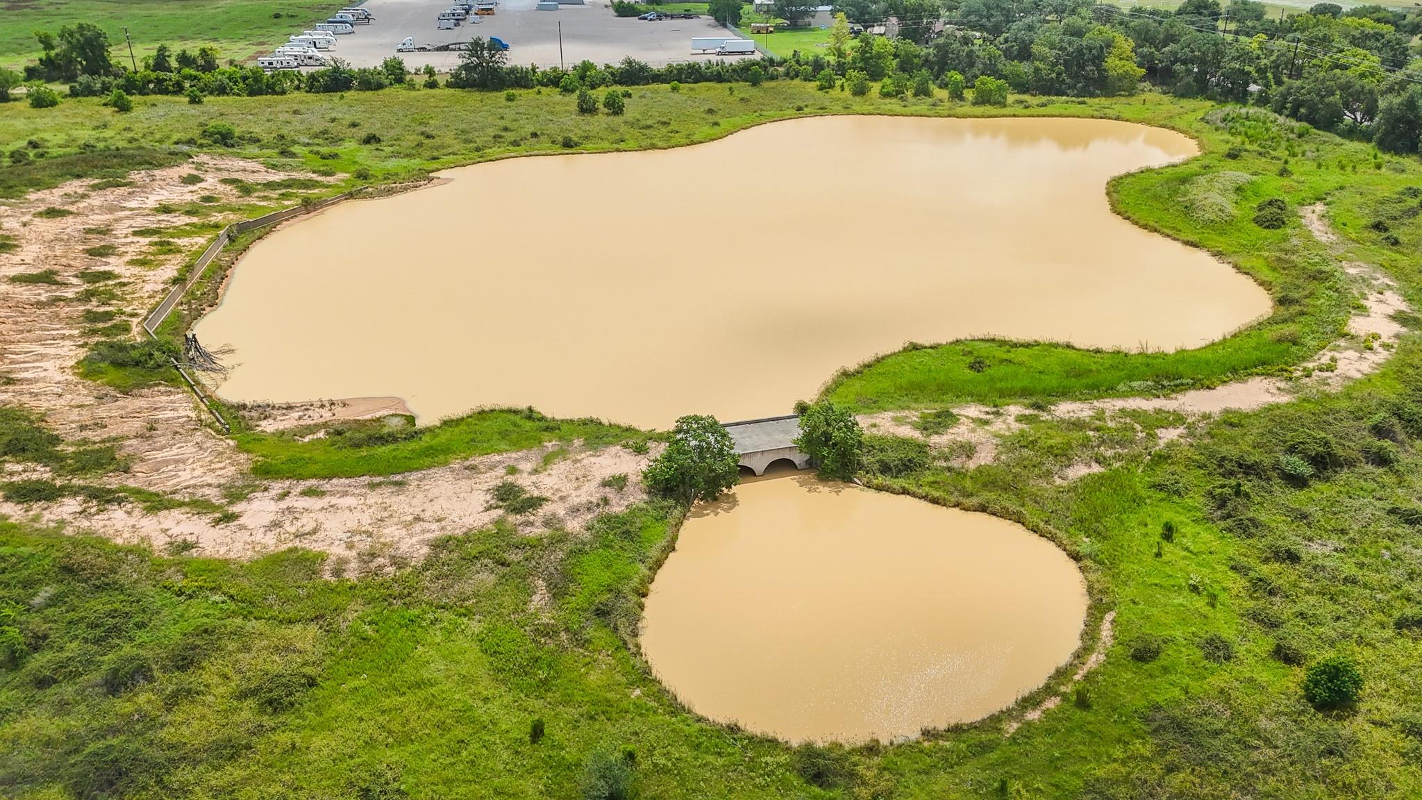 Aerial view of two muddy ponds surrounded by grassy land, with a culvert between them.