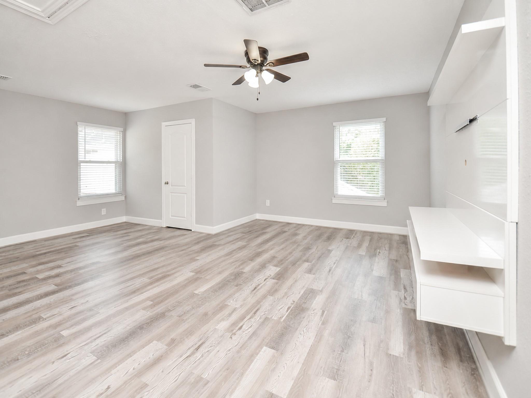 Empty room with gray plank flooring, two windows, a ceiling fan, and white wall-mounted media shelving.