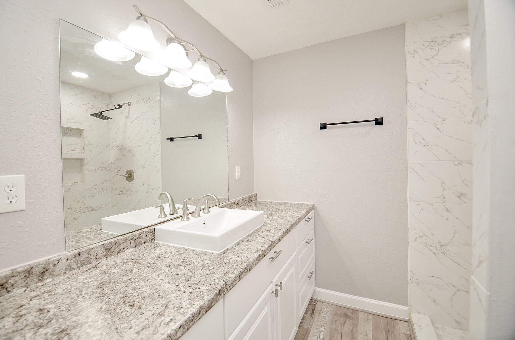 Bathroom vanity with granite countertop and vessel sink beside a marble-look tiled shower.