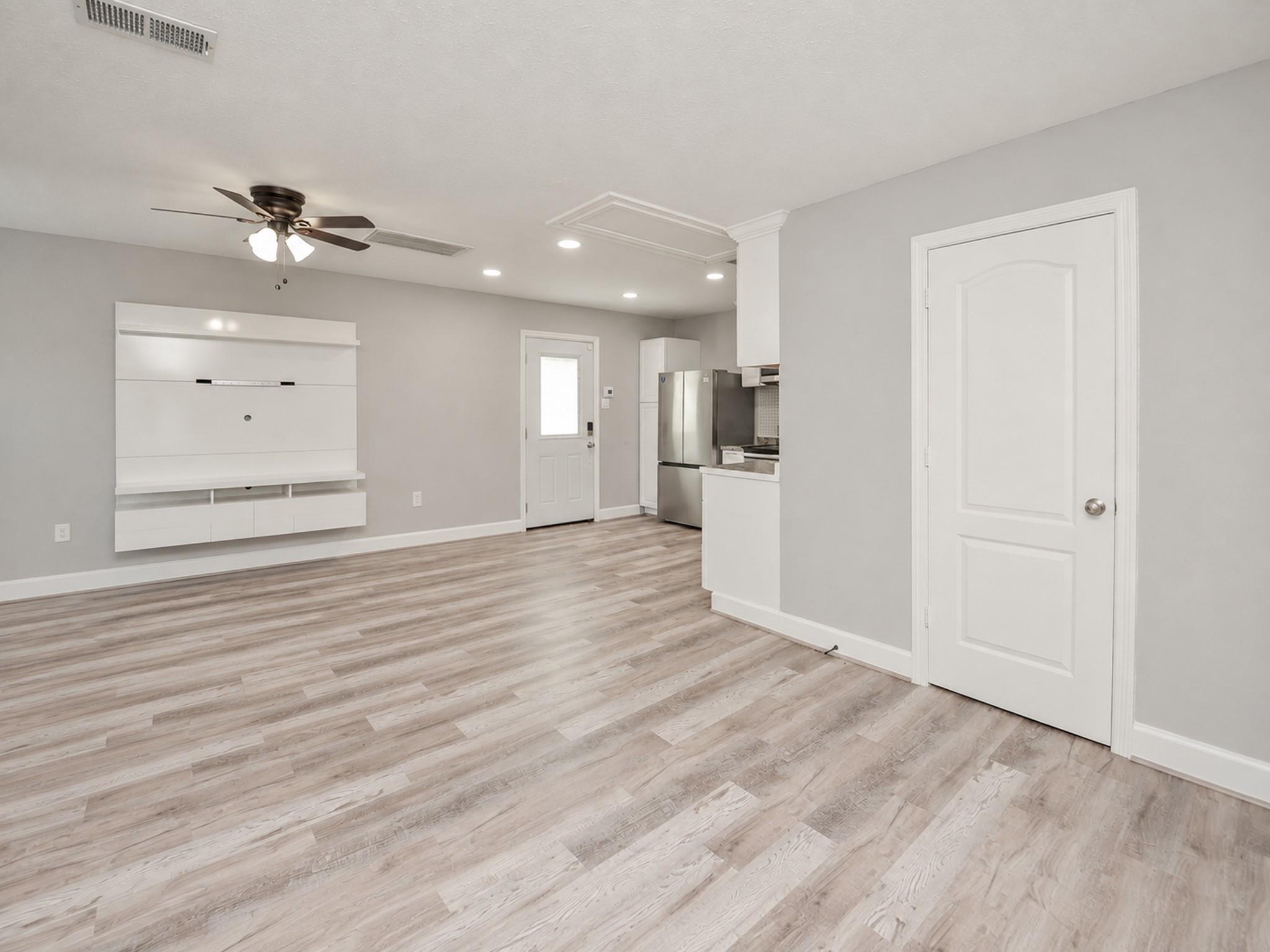 Empty living room with wood-look floors, built-in media cabinet, ceiling fan, and open kitchen.