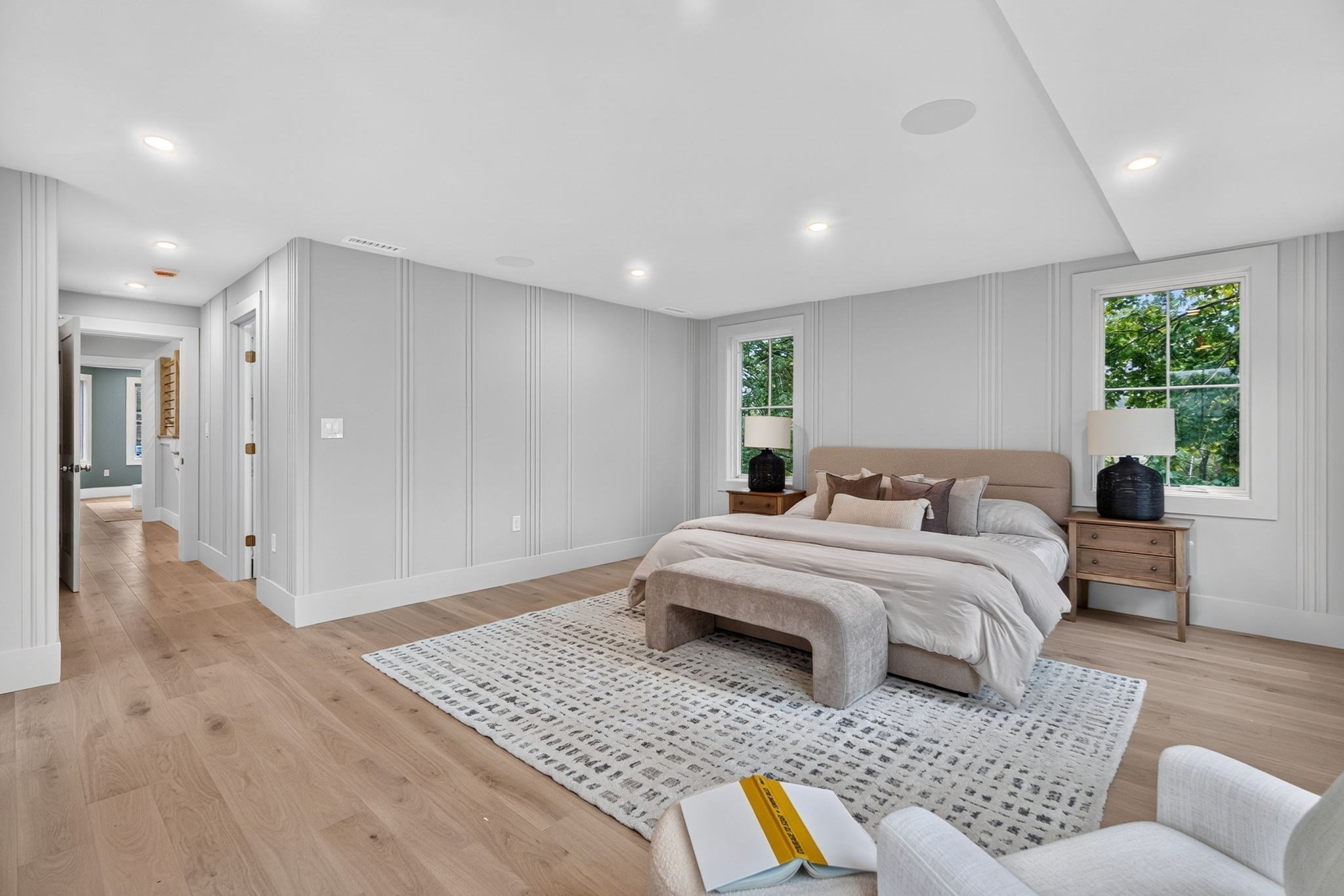 Primary bedroom with paneled gray walls, a large bed, wood floors, and two leafy window views.