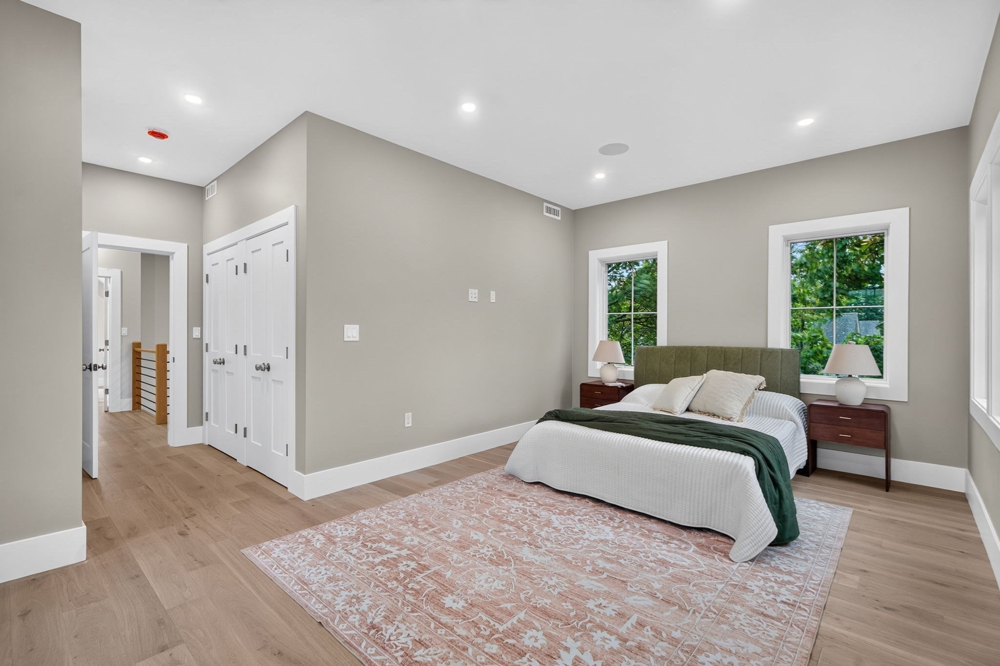 Bedroom with an olive green headboard, leafy views through two windows, and a patterned rug.