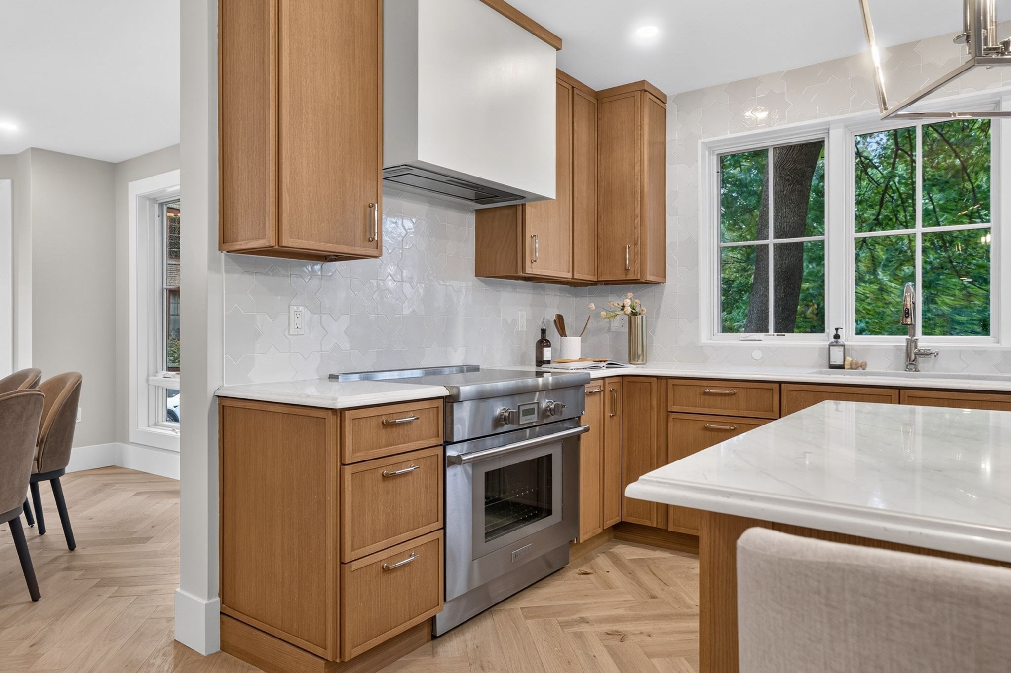 Kitchen with warm wood cabinets, stainless steel range, marble island, and large window facing leafy trees.