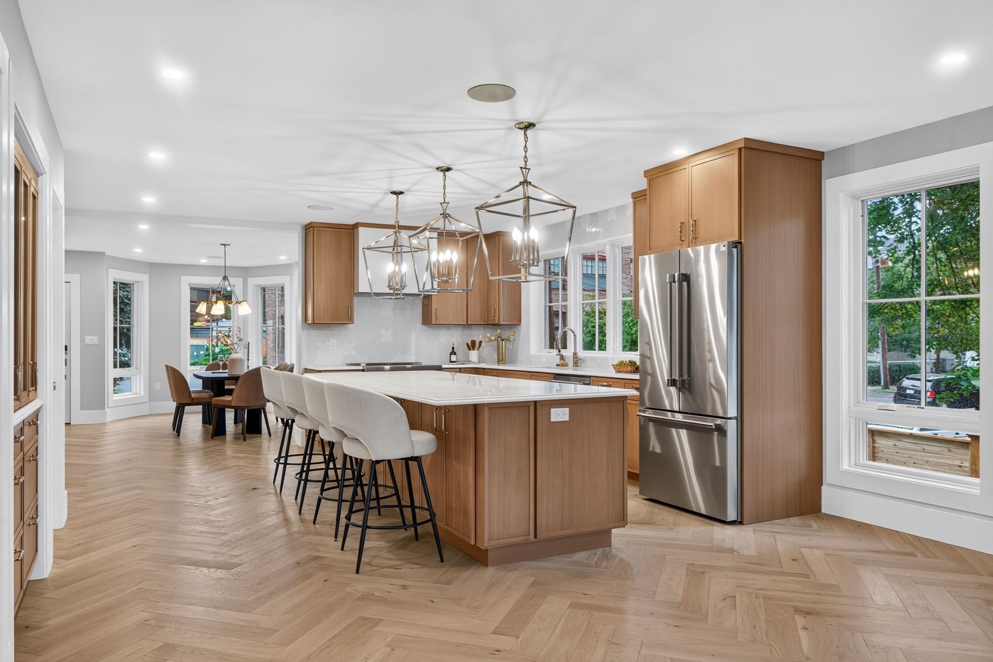 Open-plan kitchen with a wood-paneled island, white bar stools, stainless steel refrigerator, and herringbone flooring