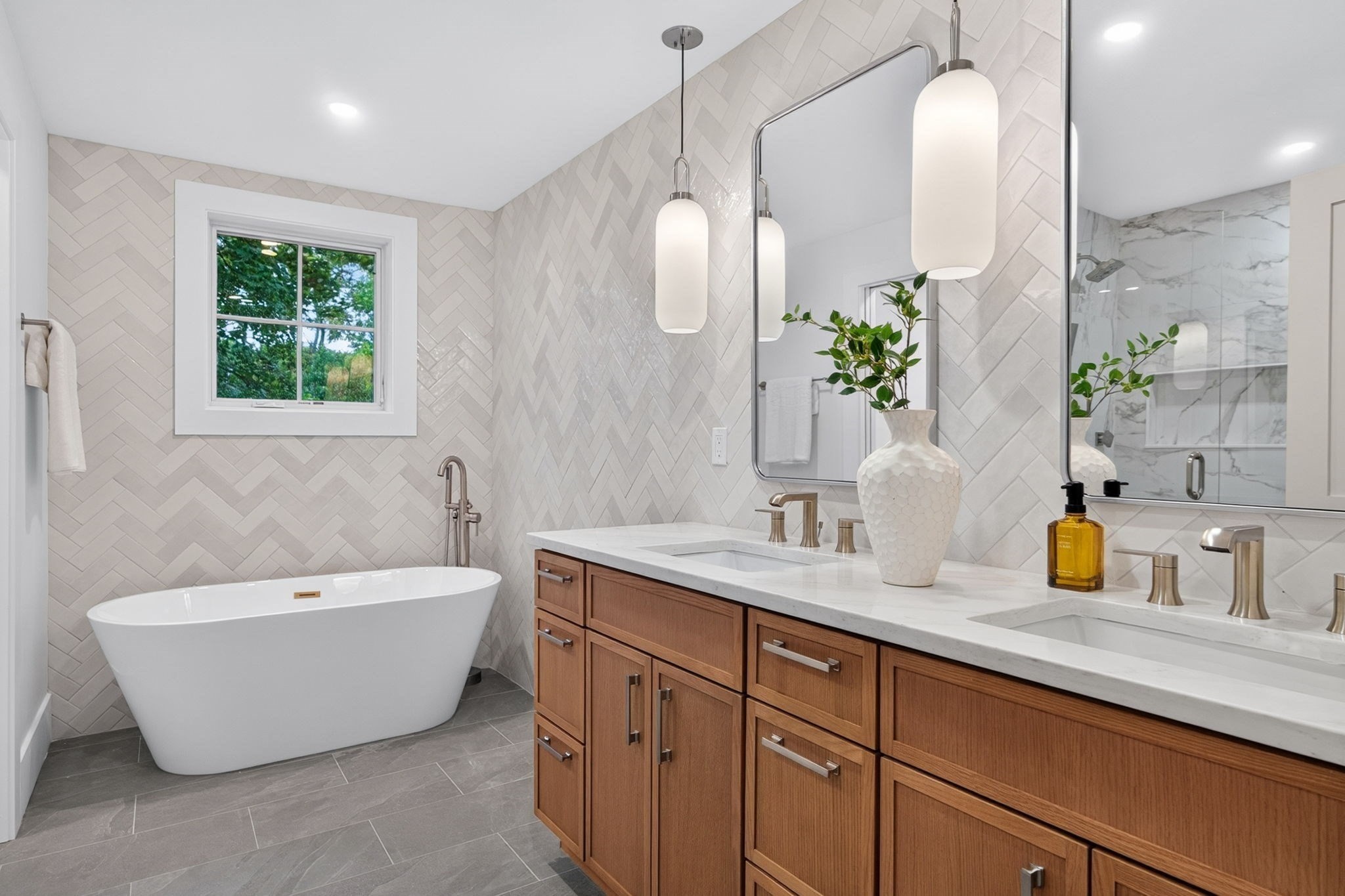 Bathroom with a freestanding tub, wood double vanity, herringbone tile, and window overlooking greenery.