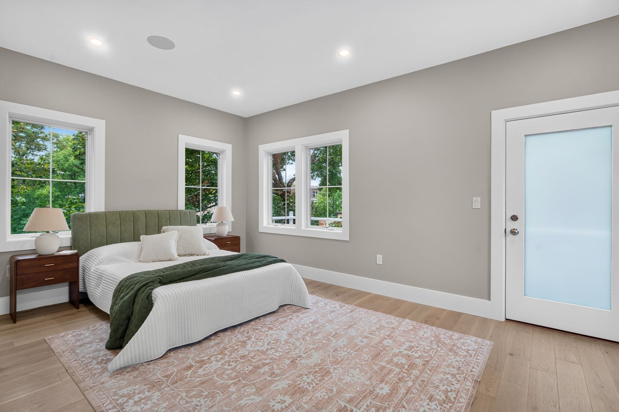 Bedroom with a green upholstered headboard, patterned rug, and windows overlooking leafy trees.