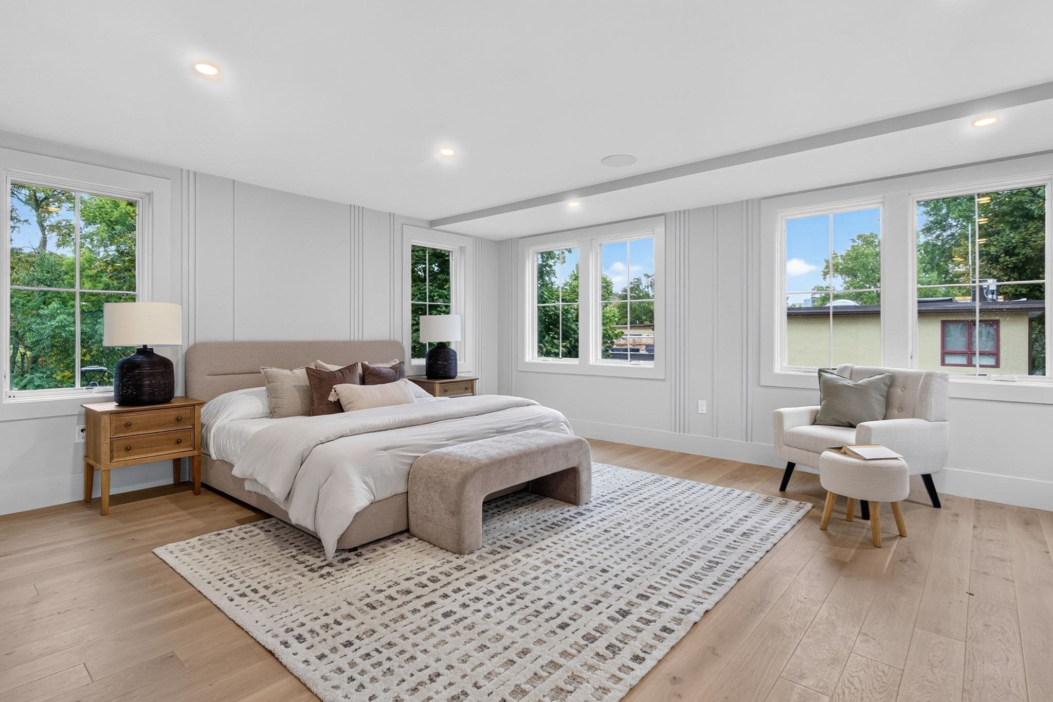 Staged bedroom with a large bed, patterned rug, and bright windows overlooking leafy trees.