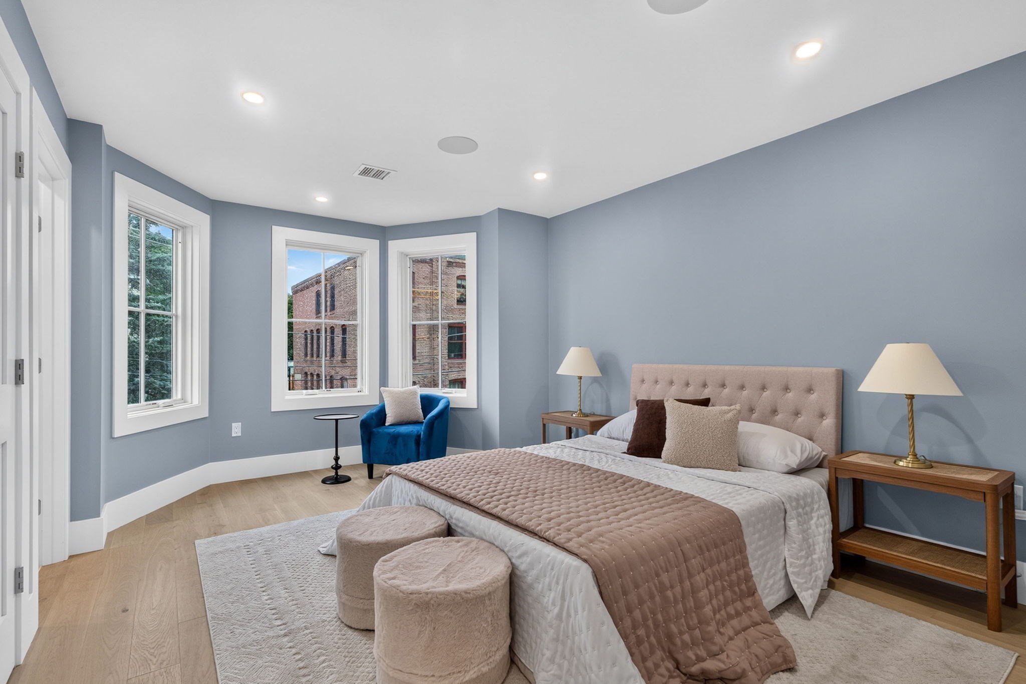 Staged bedroom with a tufted bed, blue accent chair, and three tall windows.