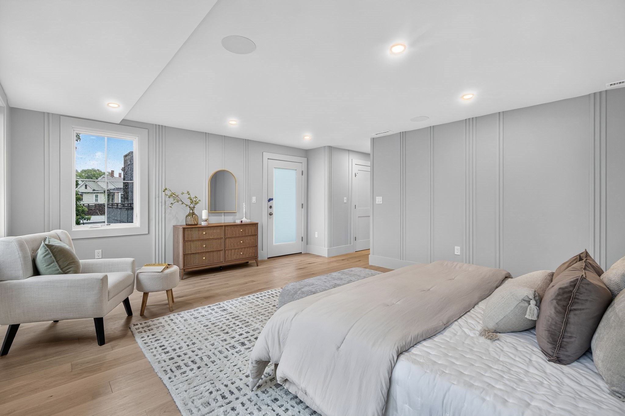 Furnished bedroom with a large bed, window-side seating, paneled walls, and light wood floors.