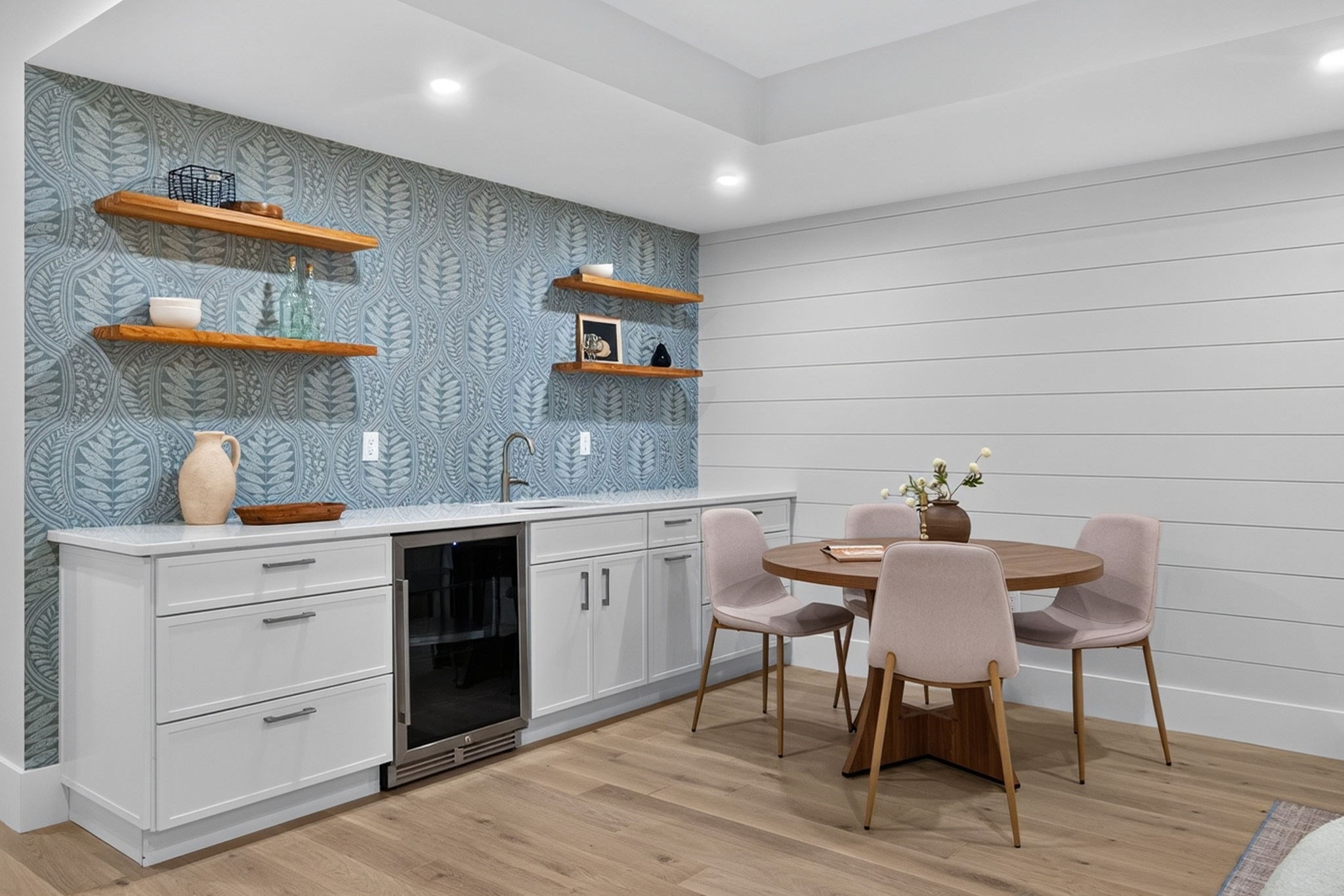 Dining nook with a round wood table, blush chairs, blue patterned wallpaper, and a built-in beverage station.