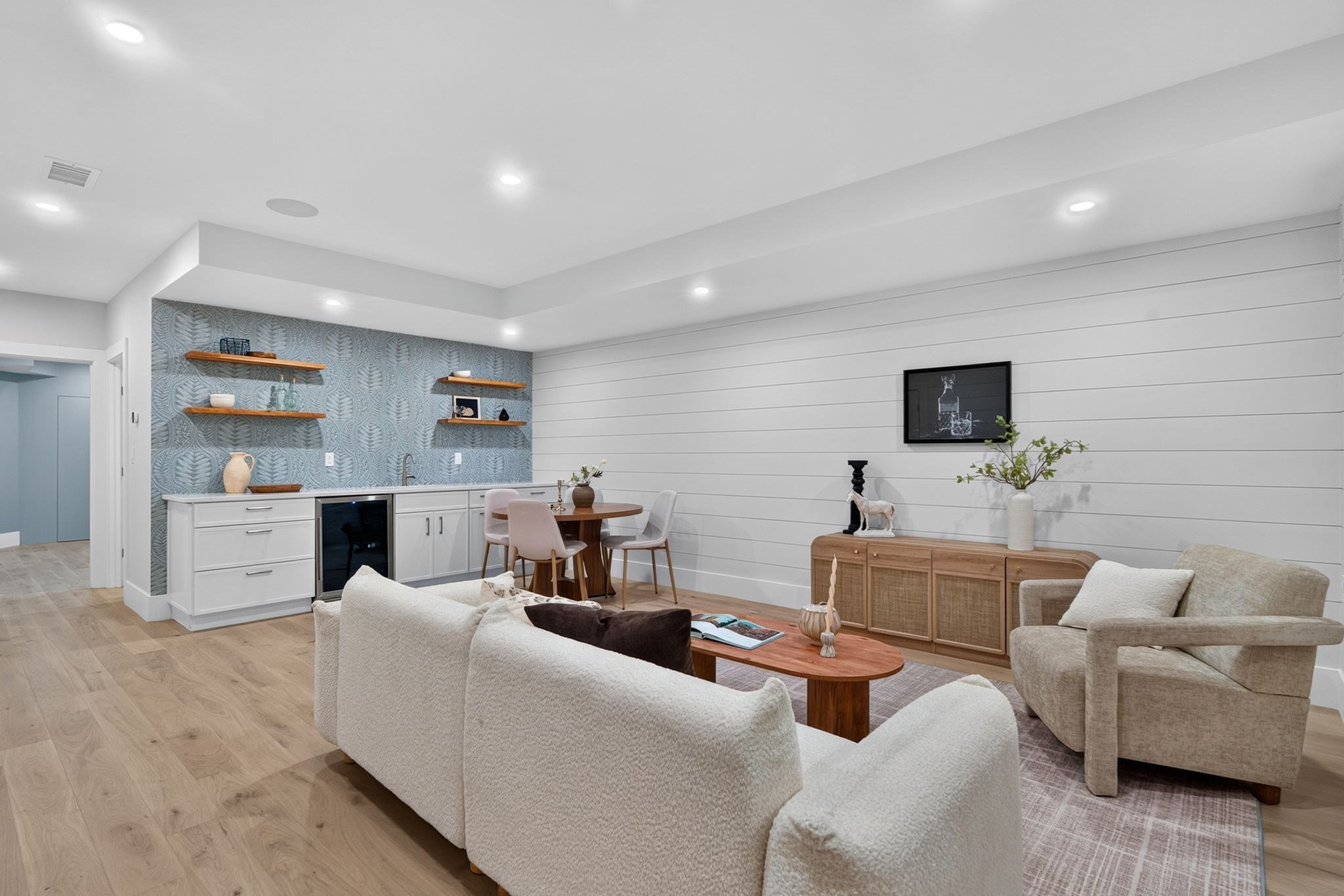 Open-plan living room with cream seating, a blue patterned kitchenette backsplash, and white paneled walls.