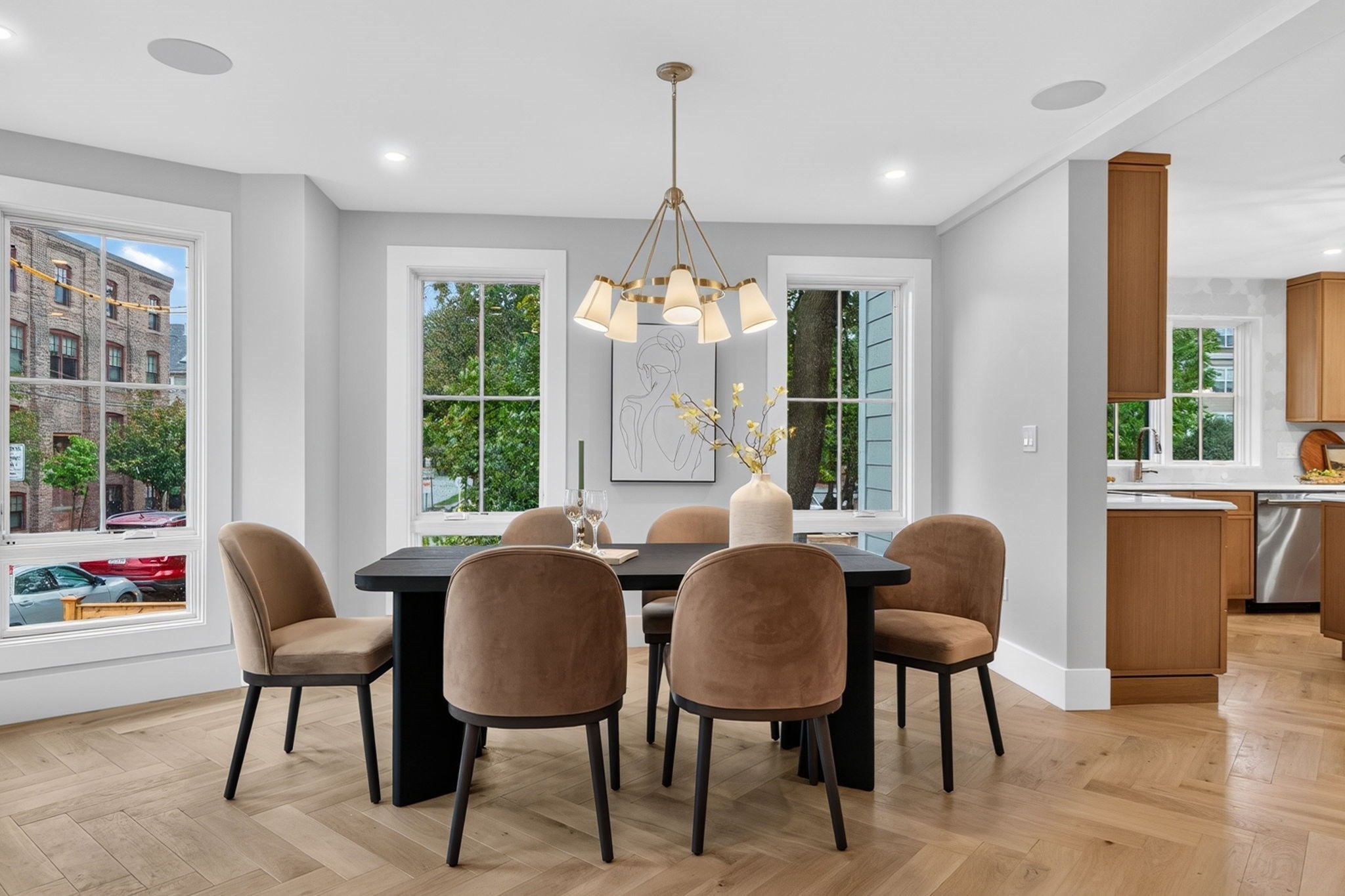 Dining room with a black table, upholstered chairs, brass chandelier, and large windows beside the kitchen.