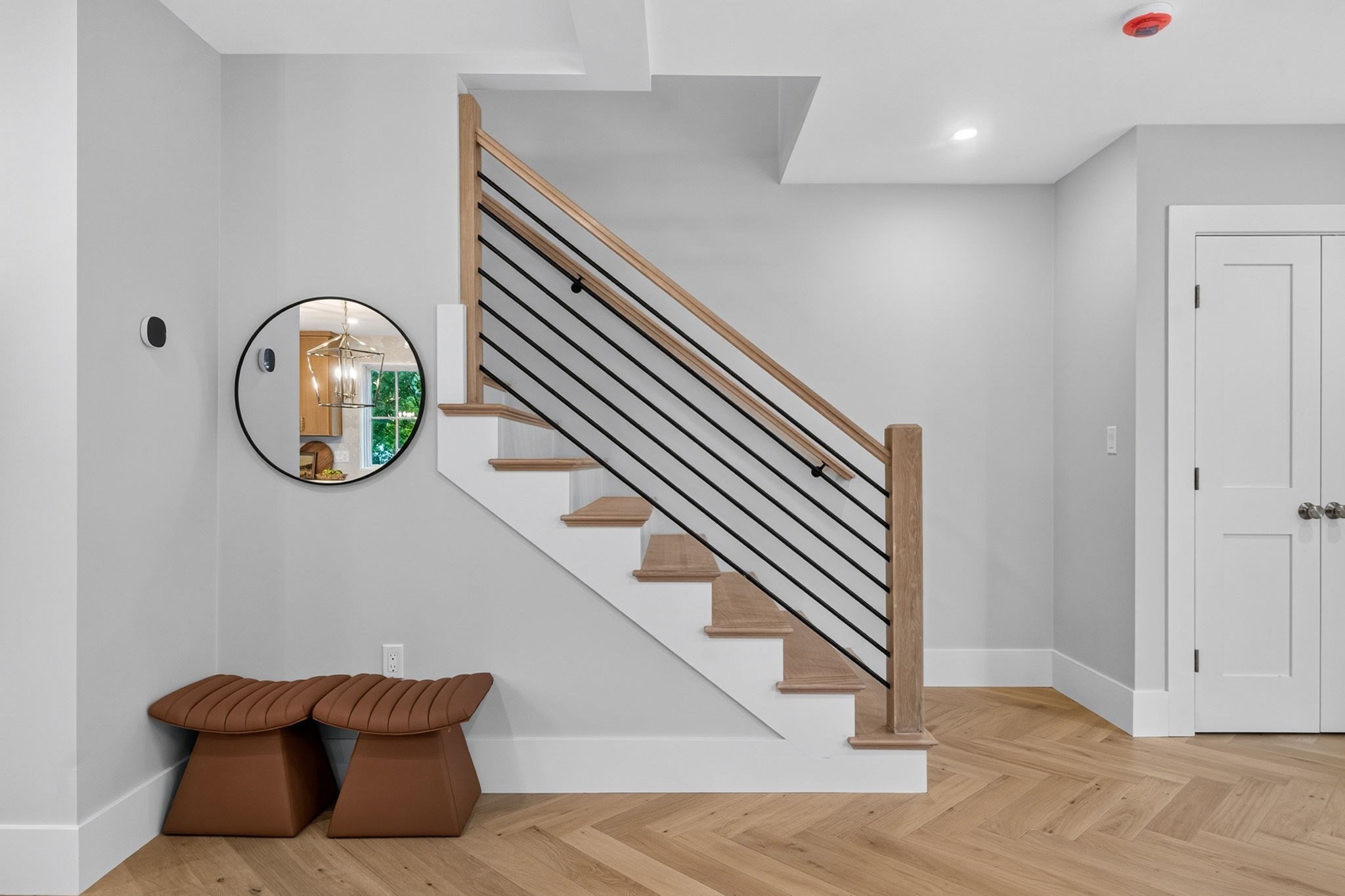 Modern entry hall with a wood staircase, black railings, round mirror, and two brown benches.