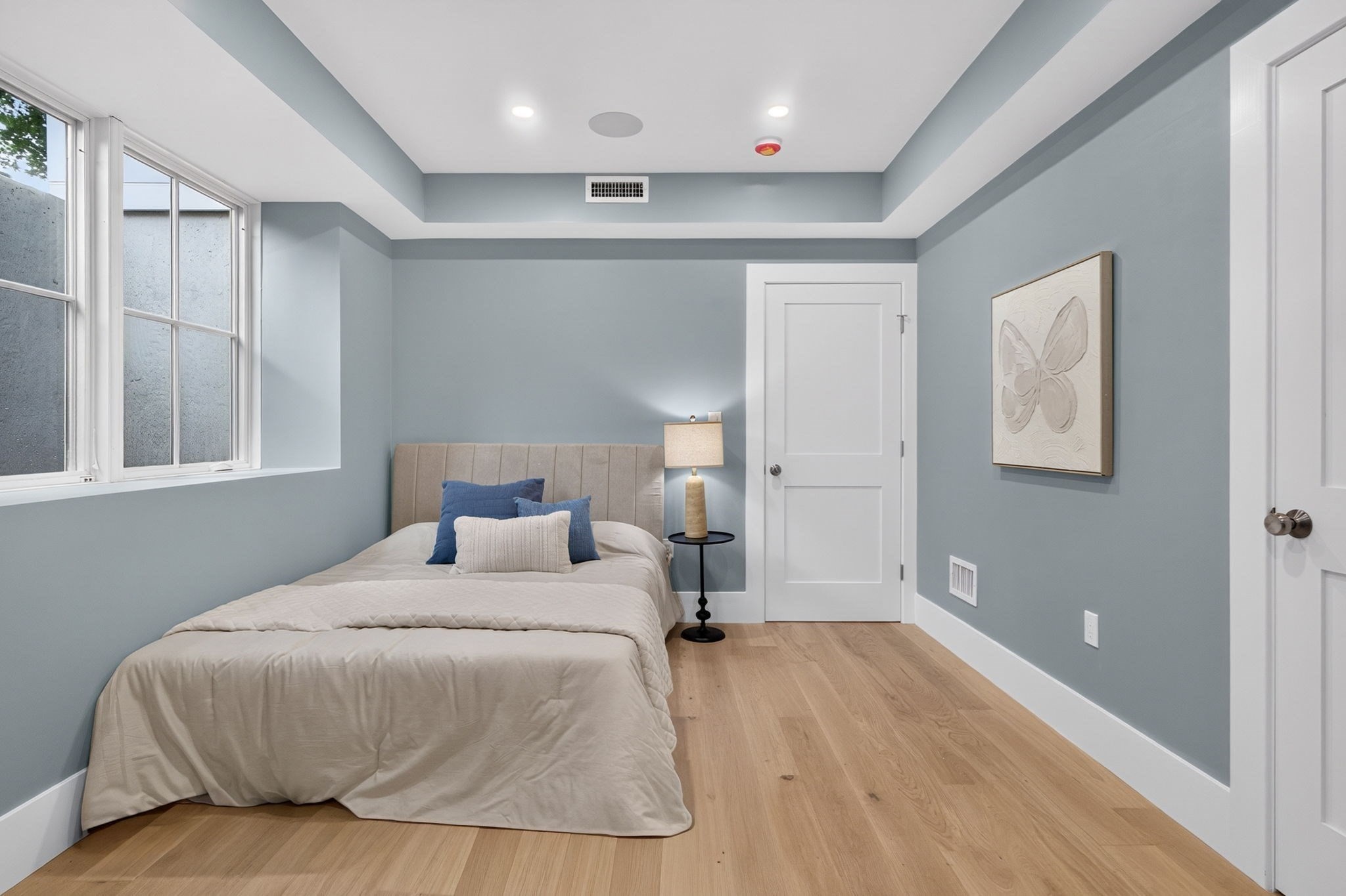 Bedroom with a beige bed, blue accent pillows, pale blue-gray walls, and large windows.