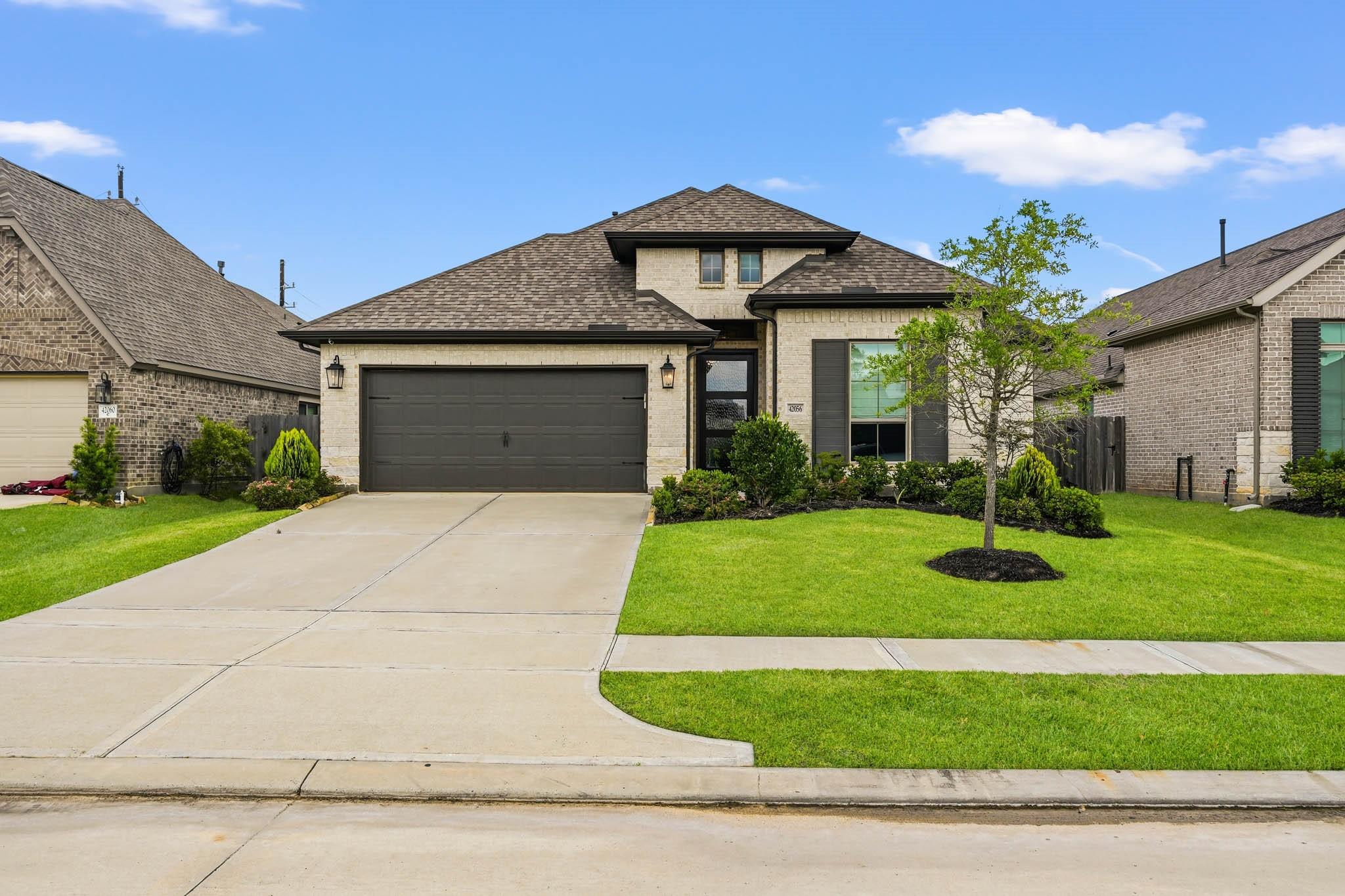 Two-story brick house with a dark double garage, landscaped lawn, and young front-yard tree.