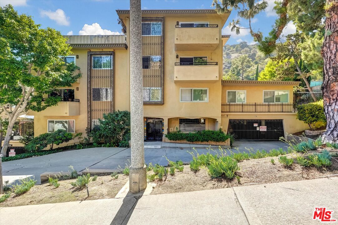 Beige multi-story residential building with balconies and gated parking, set among trees on a hillside.