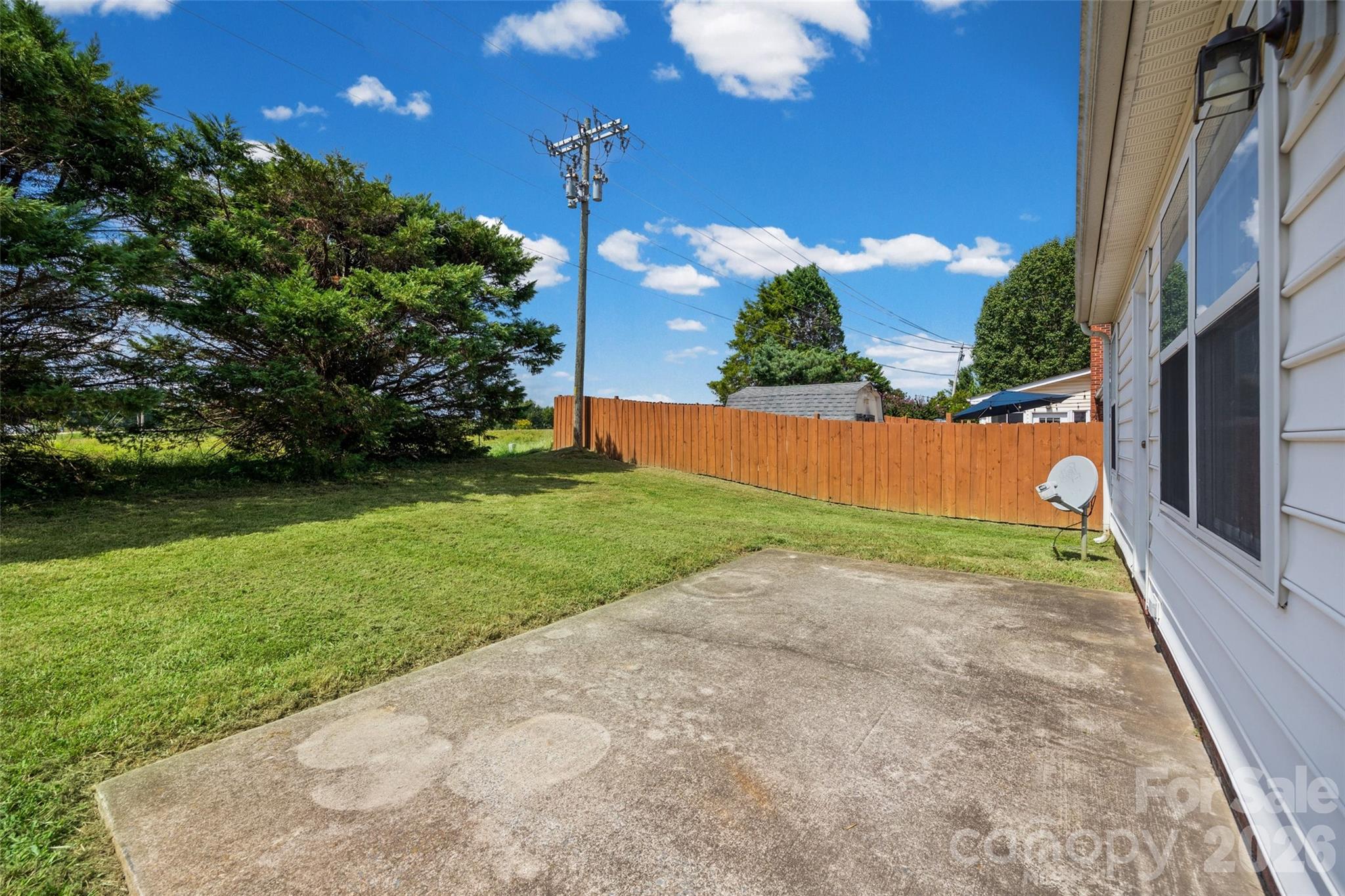 Concrete patio beside a fenced backyard with a broad lawn and mature trees.
