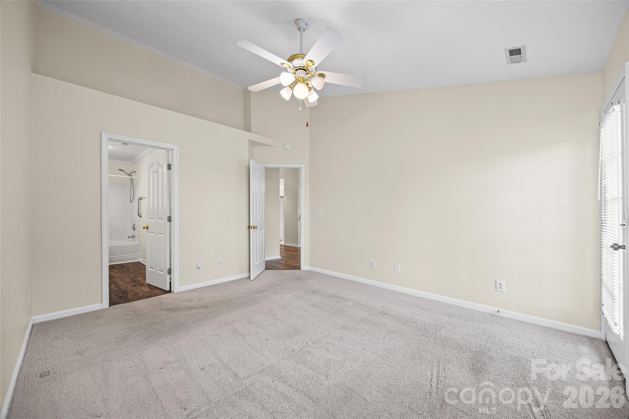 Vacant bedroom with beige carpet, ceiling fan, and open doors to a bathroom and hallway.