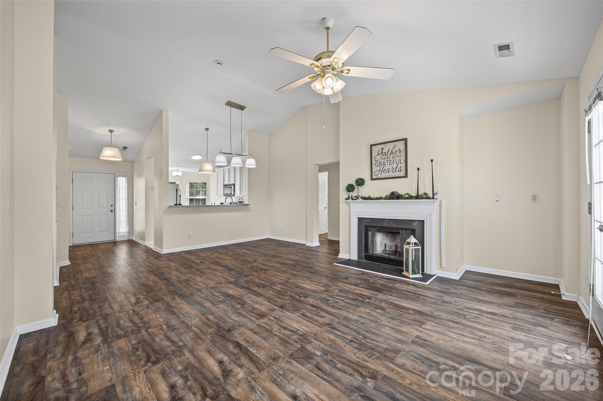 Unfurnished living room with dark plank flooring, a white fireplace, vaulted ceiling, and open kitchen view.