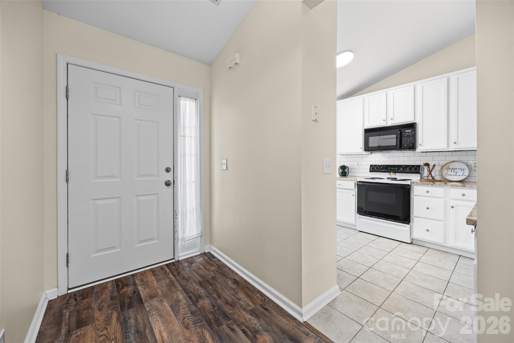 Entry foyer opening to a bright kitchen with white cabinets, black appliances, and tiled floors.