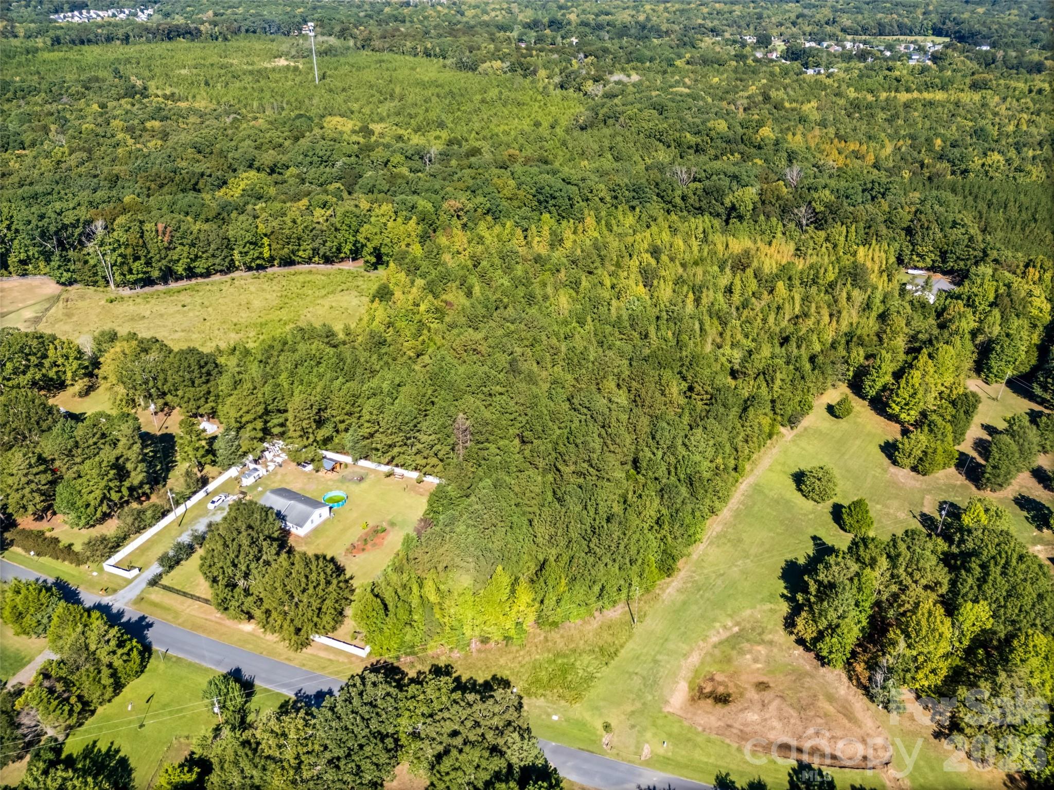 Aerial view of a wooded property with a home, above-ground pool, open fields, and surrounding roads.