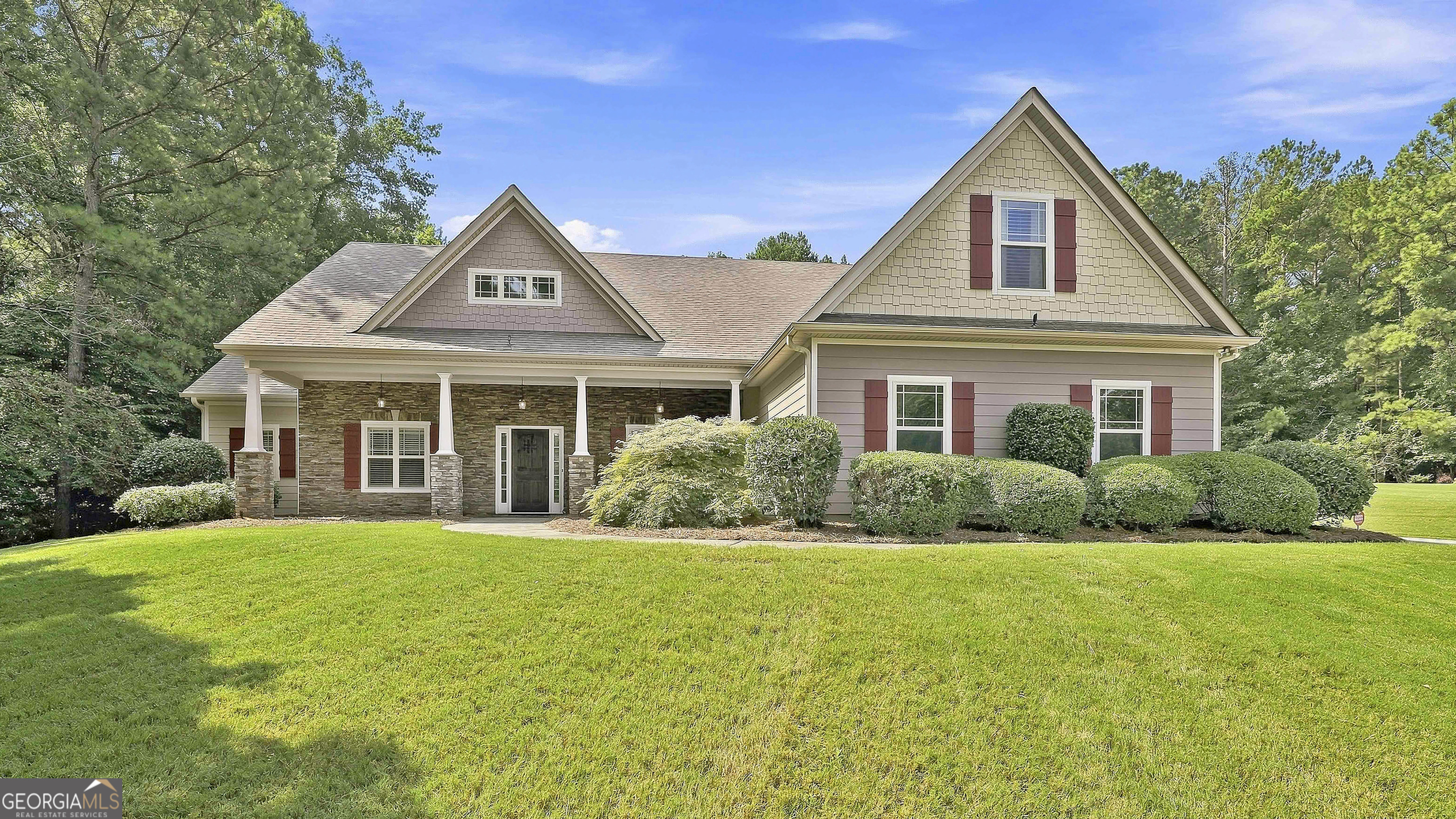 Two-story beige home with red shutters, stone porch, and a broad green front lawn.