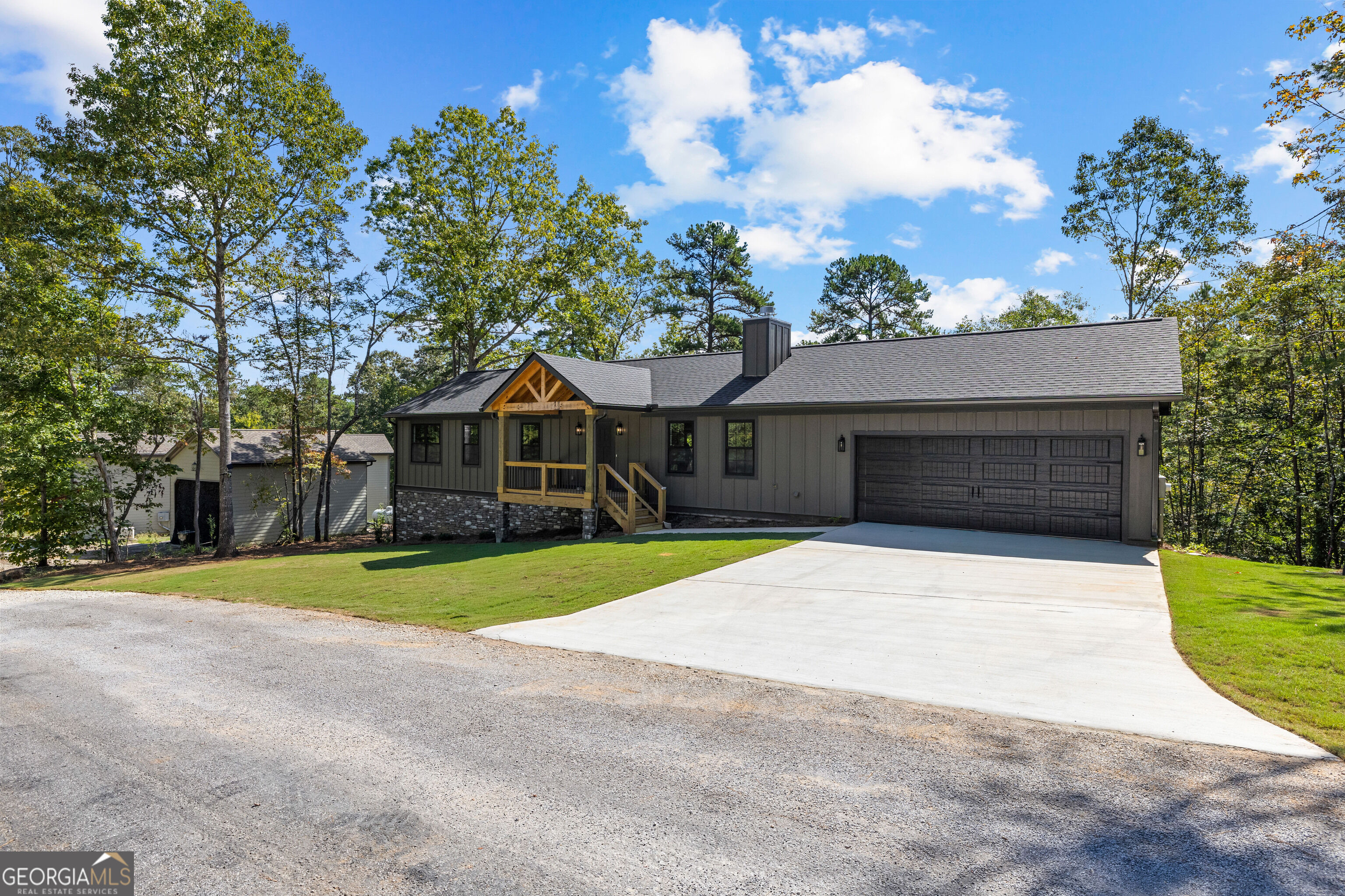 Dark gray home with a two-car garage, timber-trimmed porch, and broad concrete driveway among mature trees.