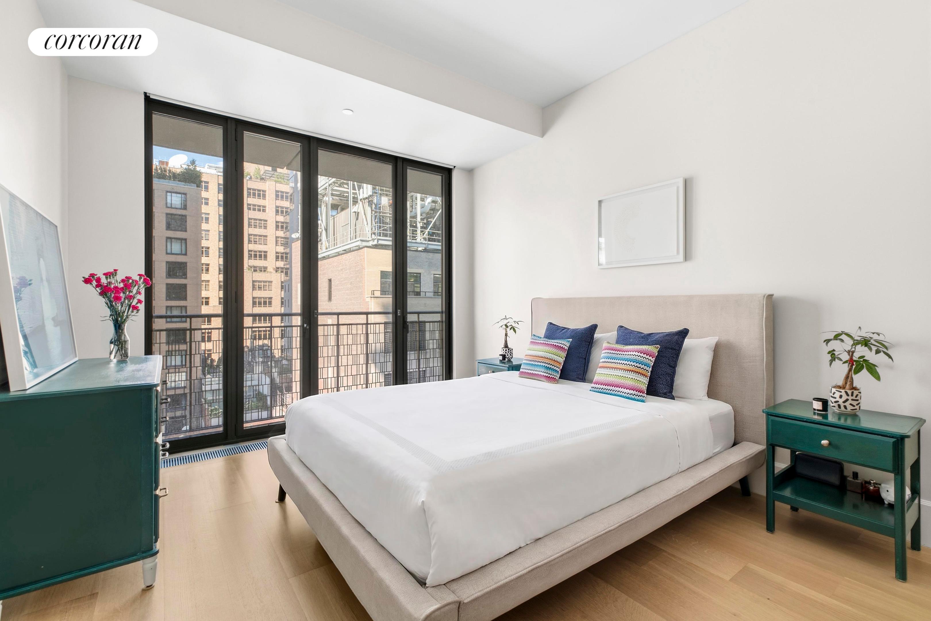 Furnished bedroom with floor-to-ceiling balcony windows and colorful pillows on a white bed.