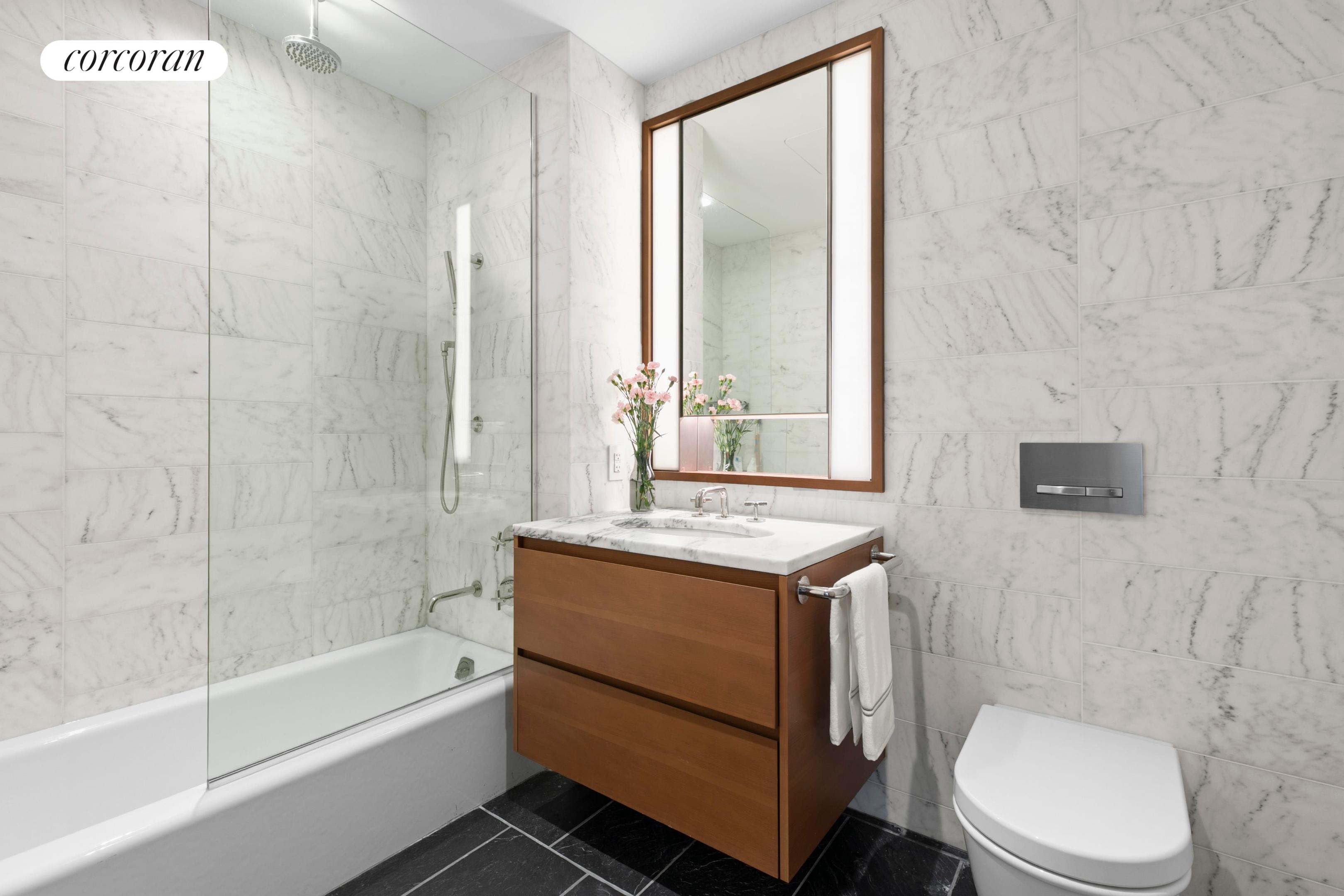 Marble bathroom with a glass-enclosed tub, wood vanity, large mirror, and toilet.