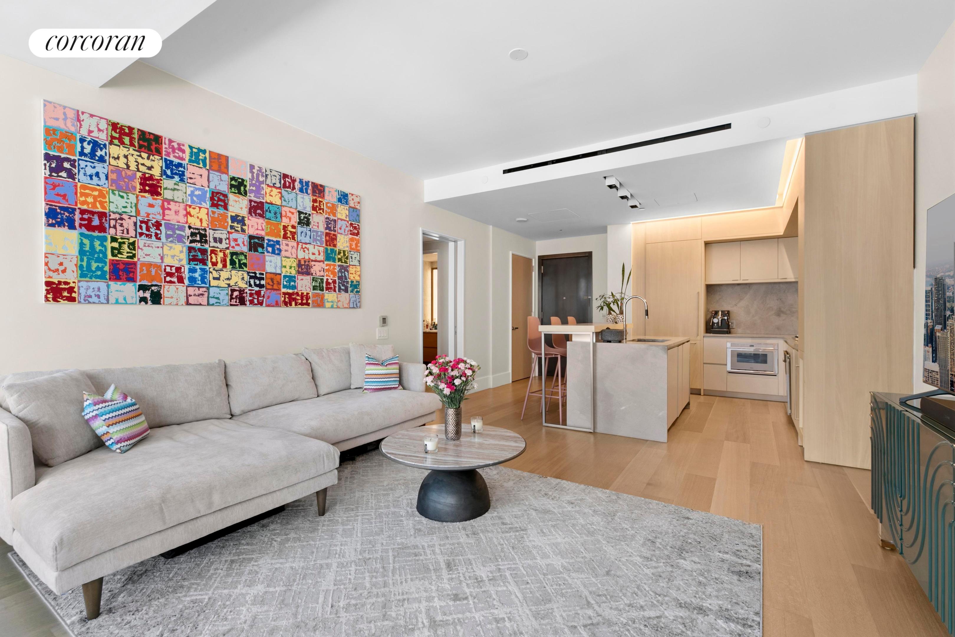 Open-plan living room and kitchen with a gray sectional, colorful wall art, and light wood cabinetry.