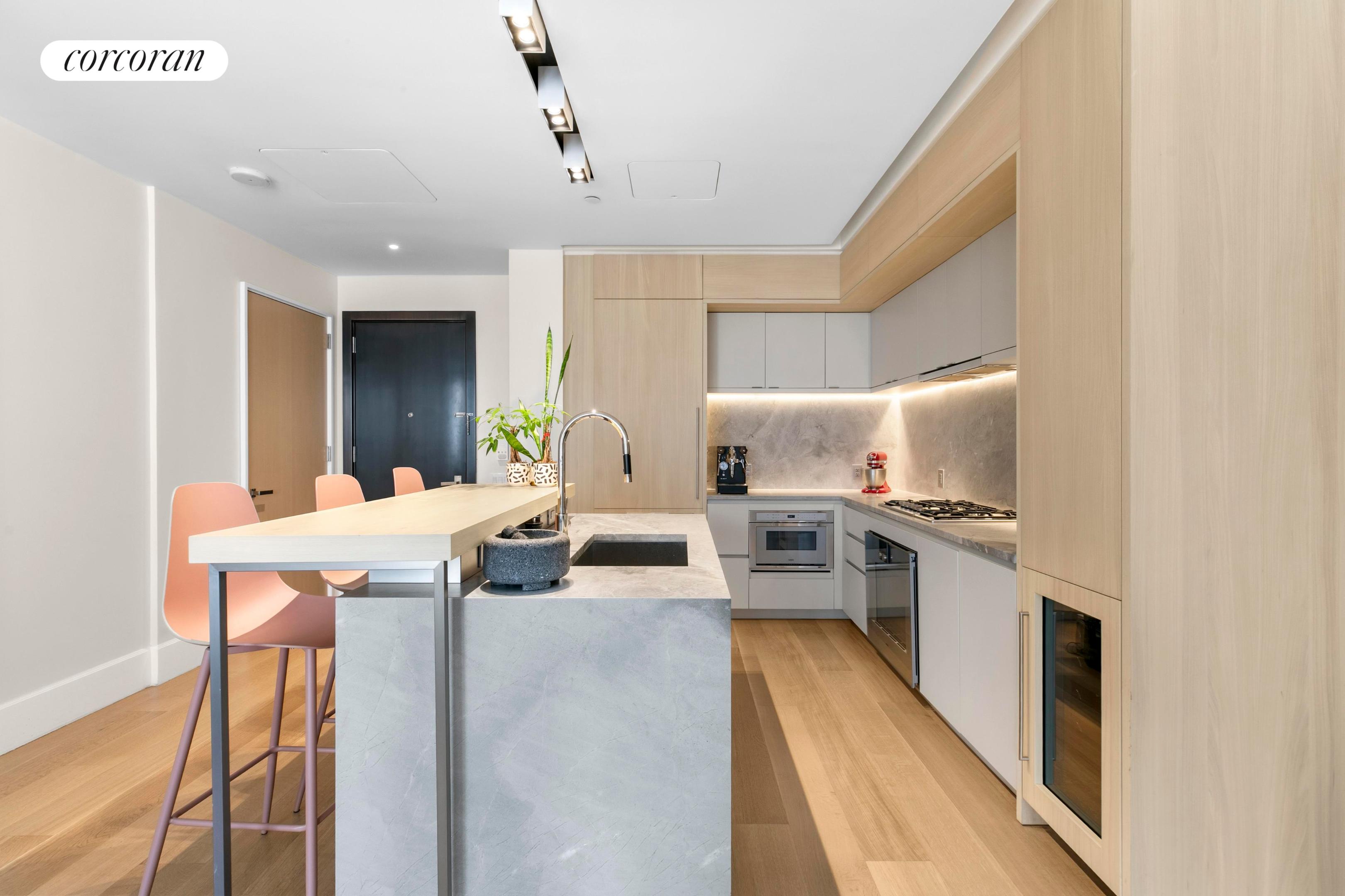 Modern kitchen with a large stone island, pale wood cabinetry, and coral bar stools.