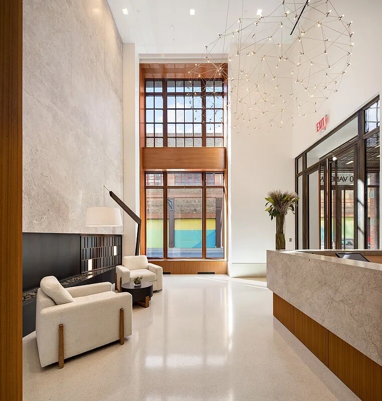 Residential lobby with tall grid windows, a branching chandelier, cream lounge chairs, and a stone reception desk.