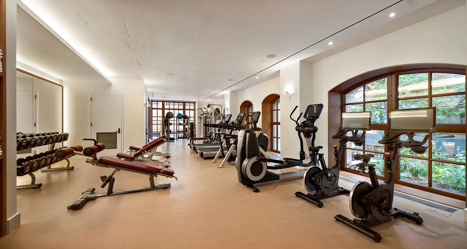 Fitness room with weight benches, cardio machines, and large arched windows overlooking greenery.