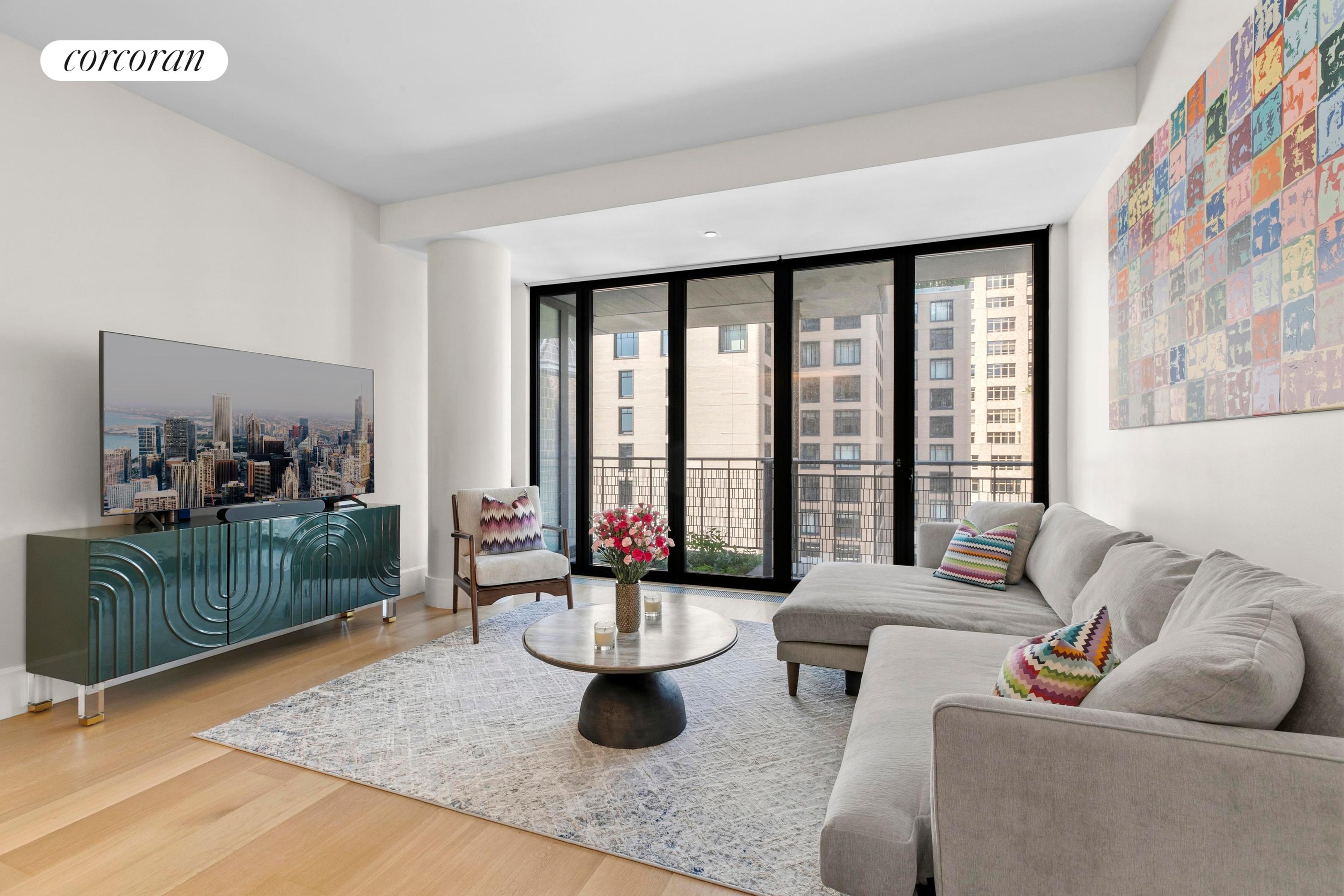 Staged living room with a gray sectional, colorful wall art, and floor-to-ceiling balcony doors.