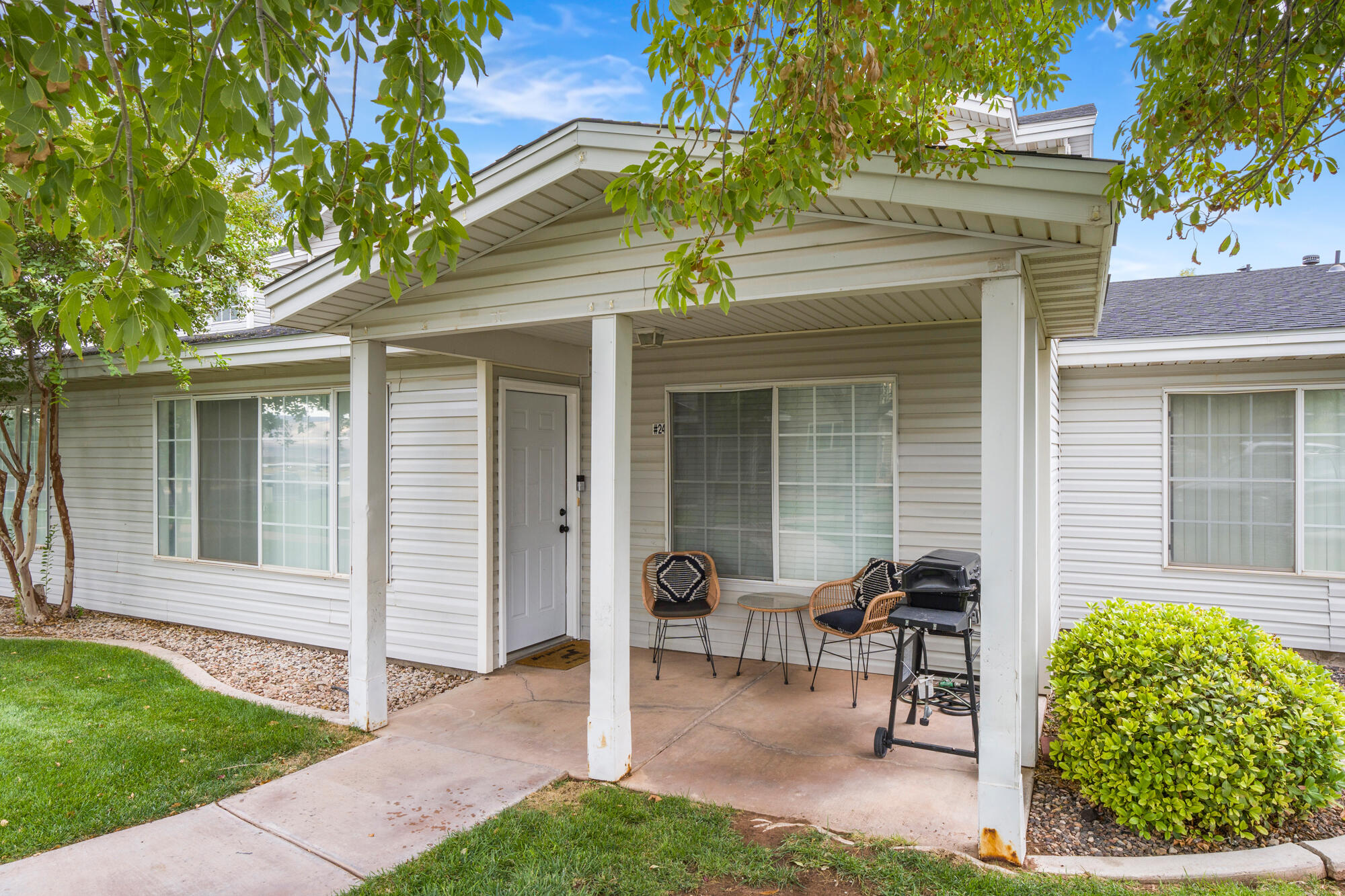 Home exterior with a covered patio, two chairs, a grill, and leafy trees.