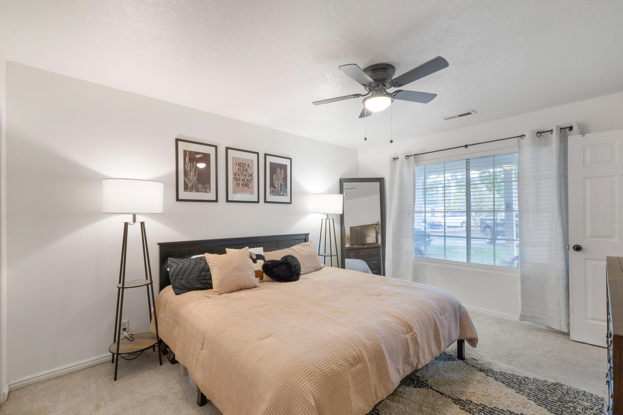 Furnished bedroom with a peach quilted bed, framed cactus prints, and a window with blinds.