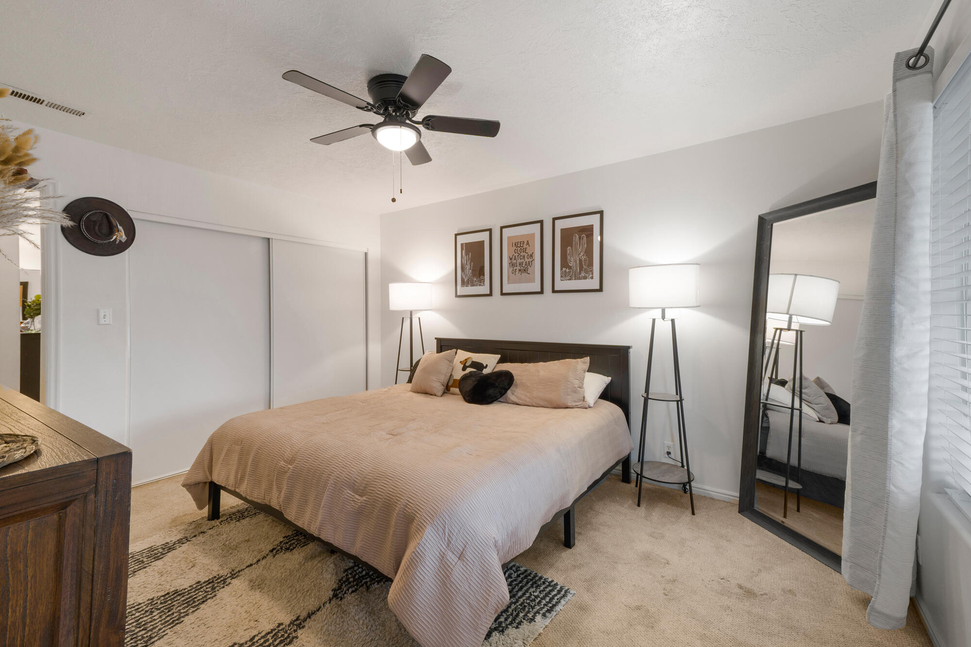 Furnished bedroom with a beige bed, matching floor lamps, framed art, and a full-length mirror.