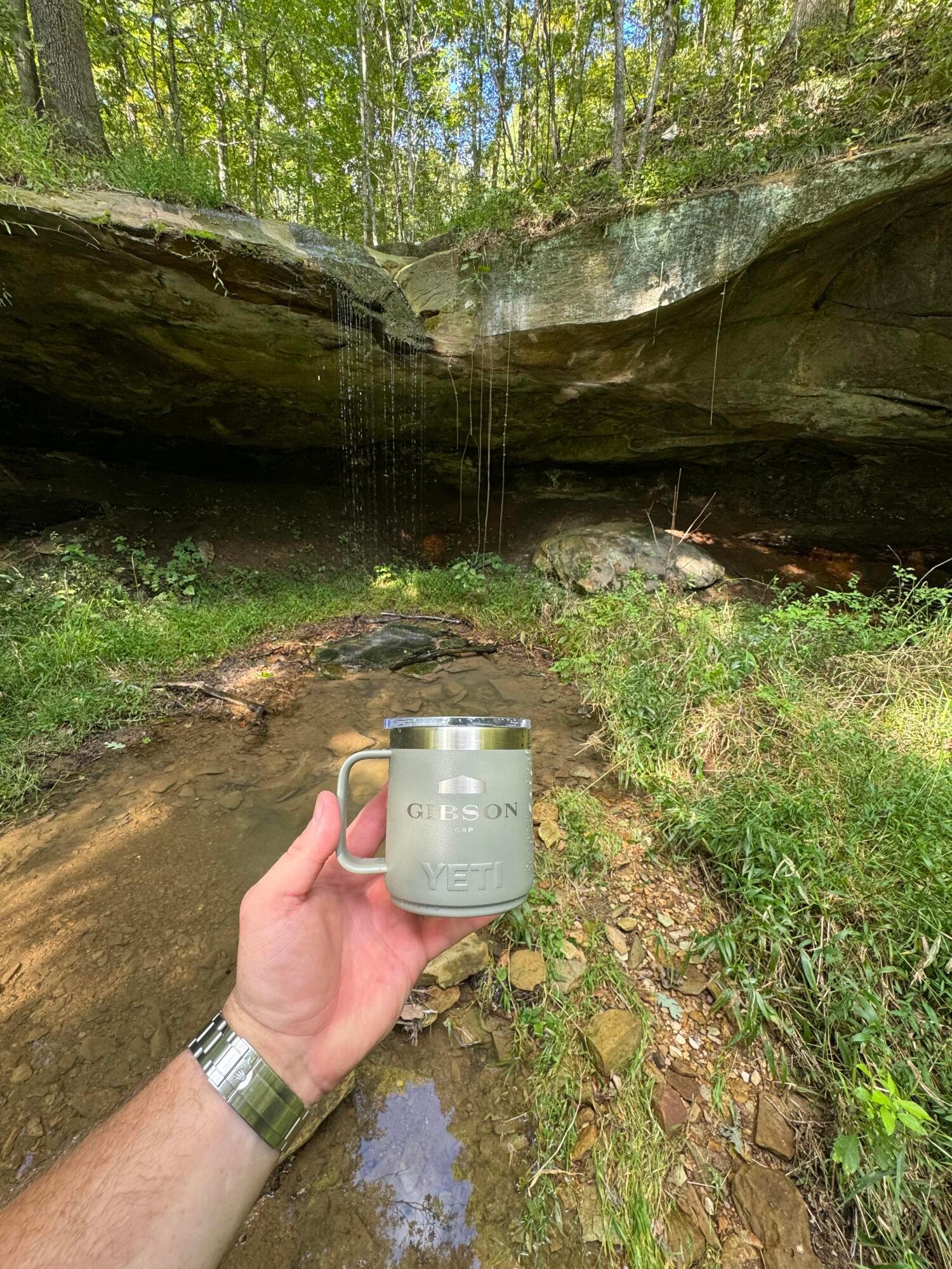 Wooded creek beneath a broad rock overhang, with a Gibson Group mug held in the foreground.