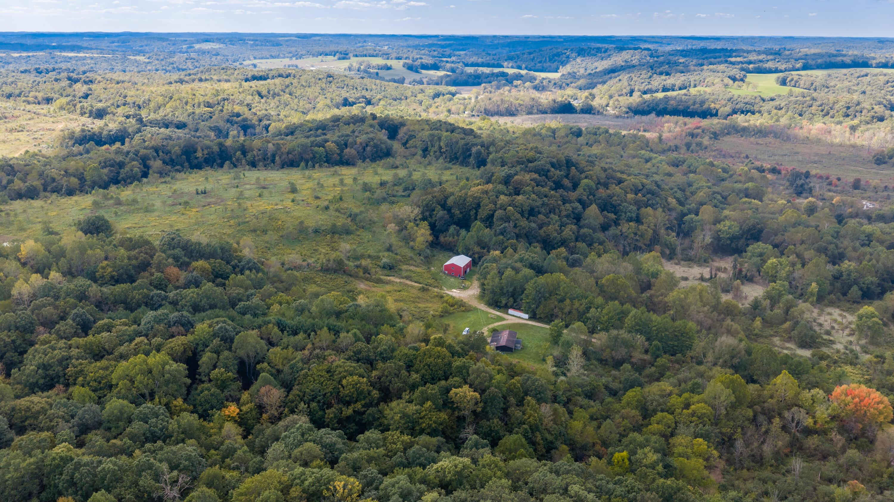 Aerial view of wooded acreage and open fields with a red barn and smaller building.