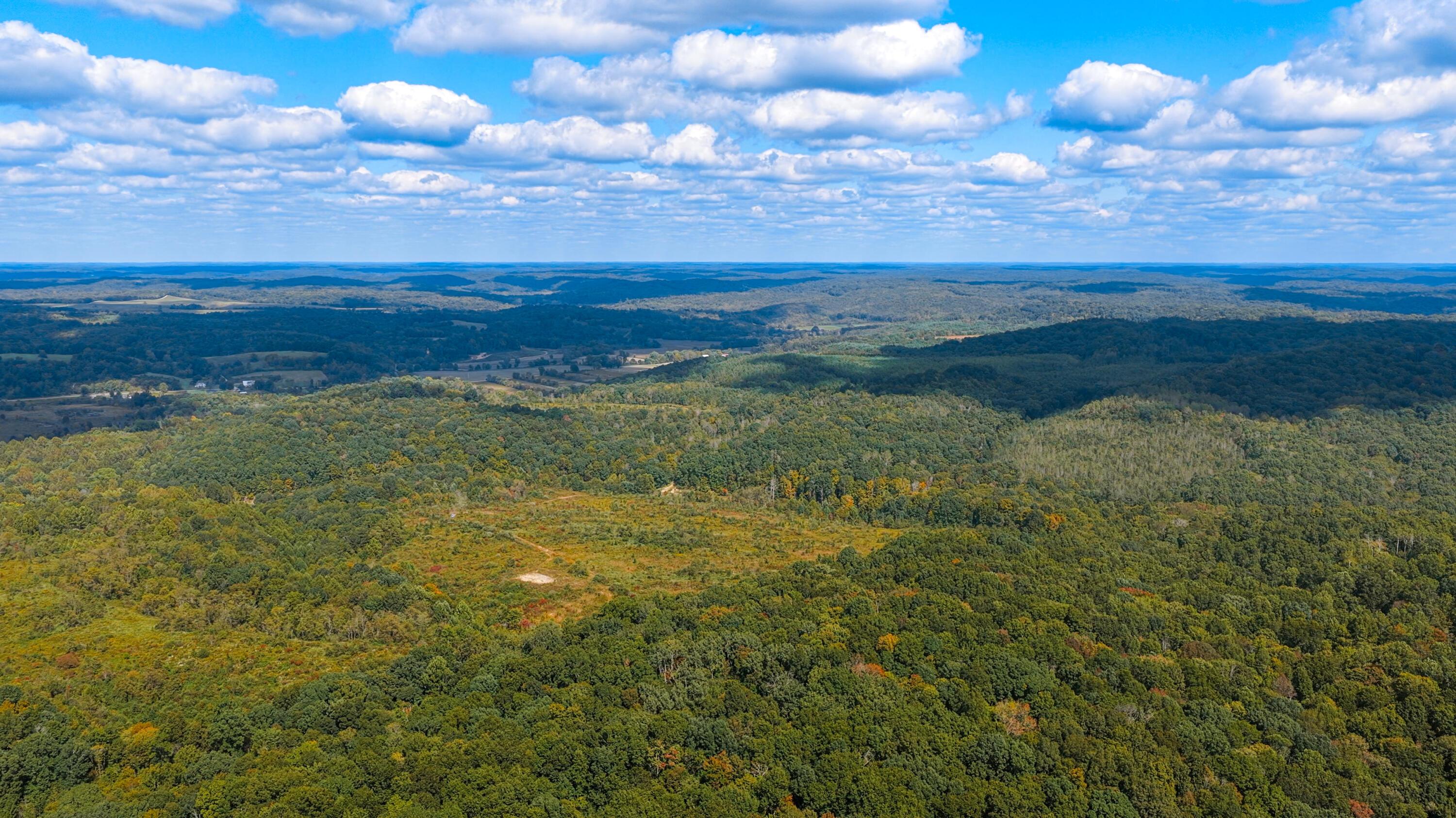 Aerial view of rolling wooded land with a small clearing and distant hills.