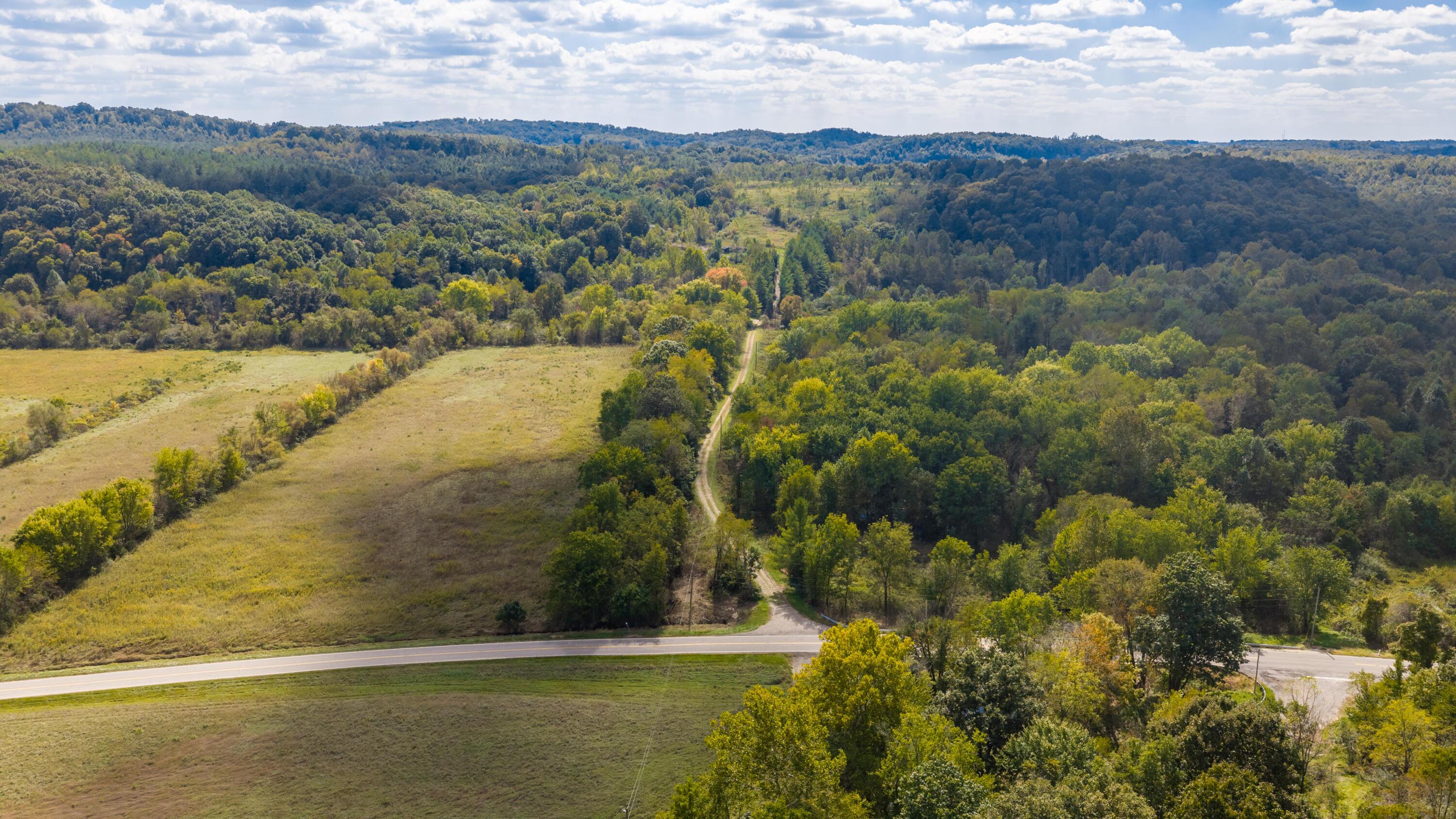 Aerial view of wooded acreage and open fields, with a dirt lane meeting a paved road.