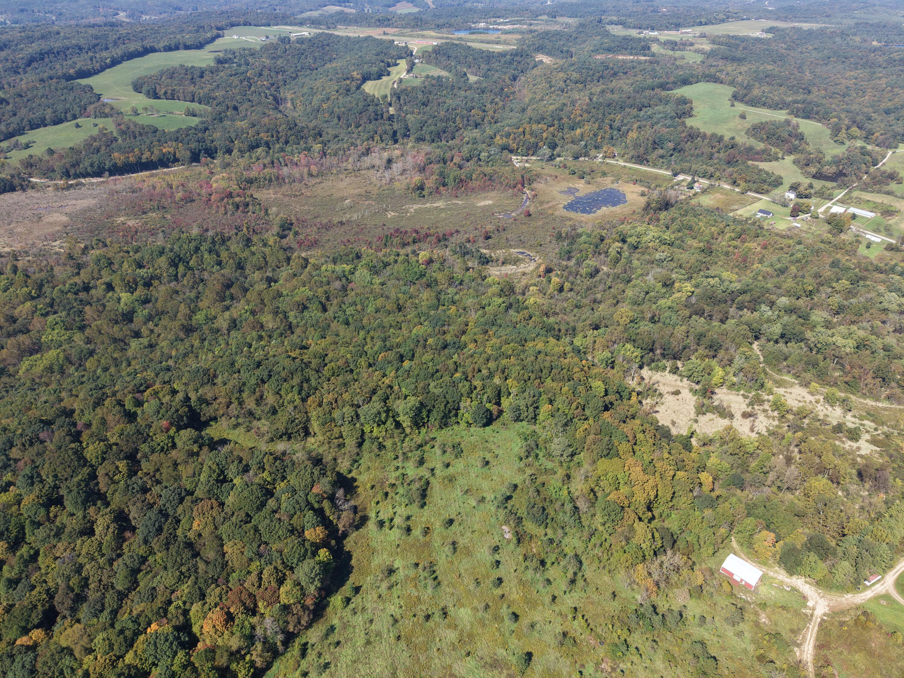 Aerial view of wooded rural land with grassy clearings, a pond, and a red-and-white barn.