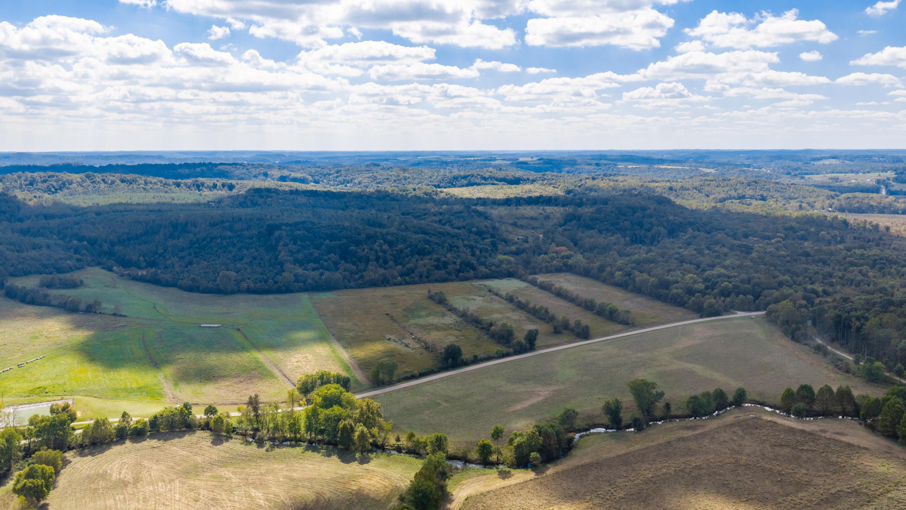 Aerial view of rural fields and wooded hills, crossed by a winding road and small creek.