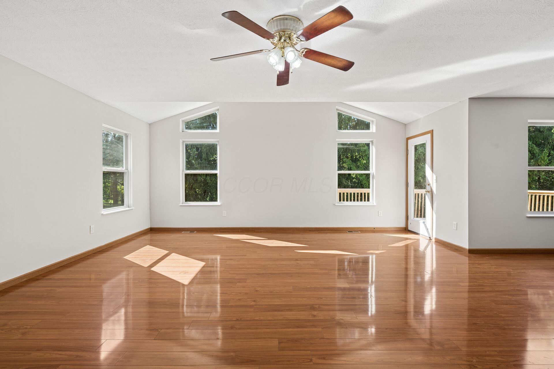 Empty living room with polished hardwood floors, vaulted ceiling, ceiling fan, and a glass door to the deck.