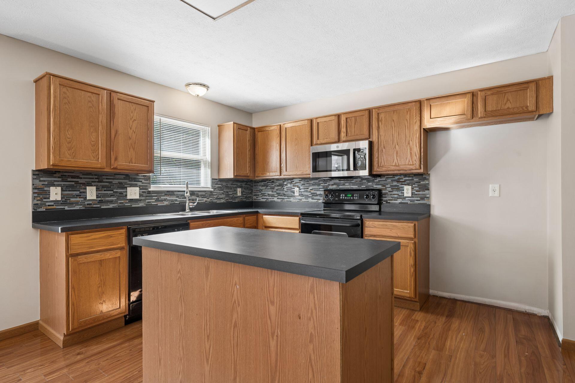 Kitchen with wood cabinetry, dark countertops, central island, and multicolored tile backsplash.