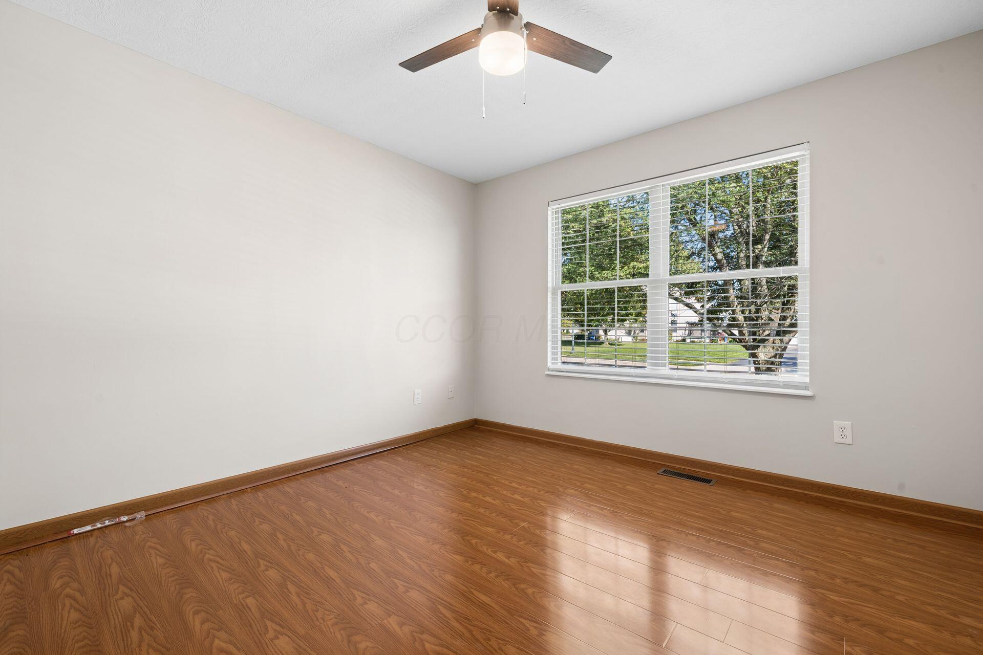 Empty room with wood floors, a ceiling fan, and a large window overlooking trees and lawn.