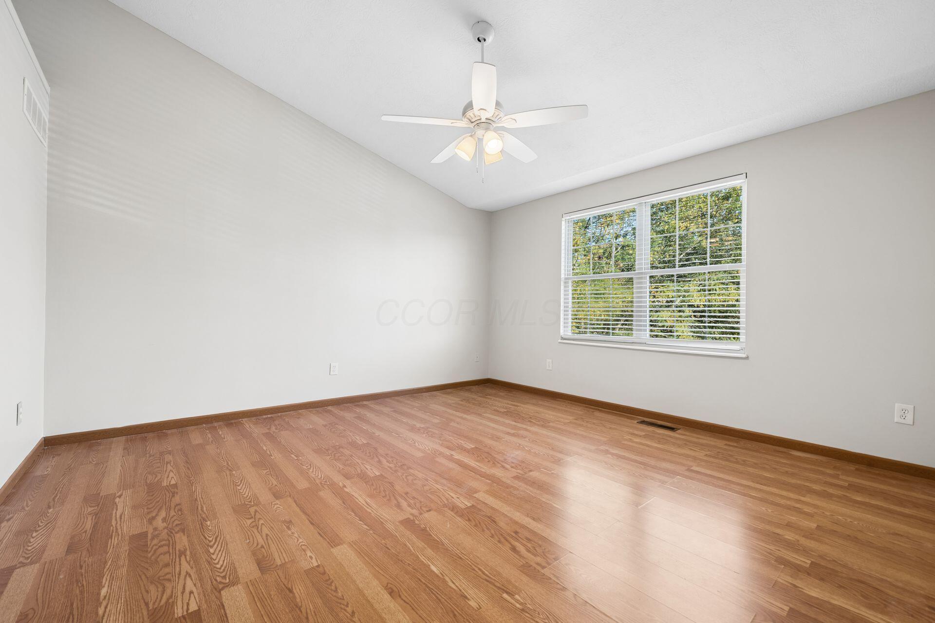 Empty bedroom with hardwood floors, a ceiling fan, and a wide window overlooking trees.