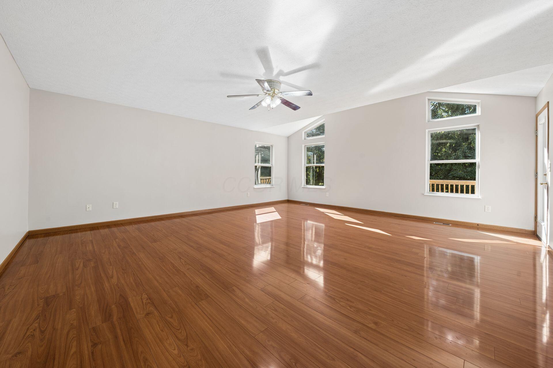 Empty living room with polished hardwood floors, vaulted ceiling, ceiling fan, and tall windows.