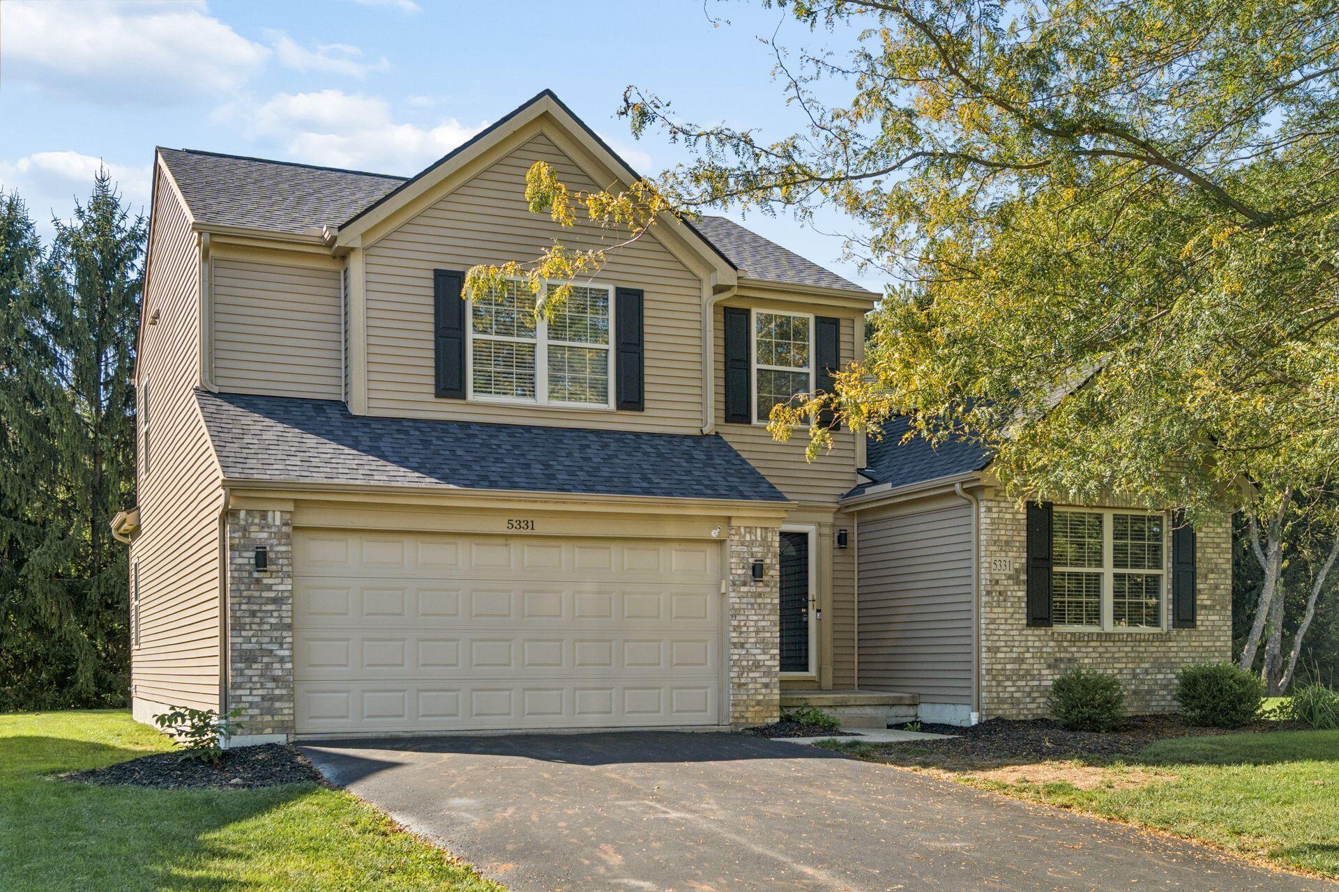 Two-story beige house with brick accents, dark shutters, and an attached double garage.