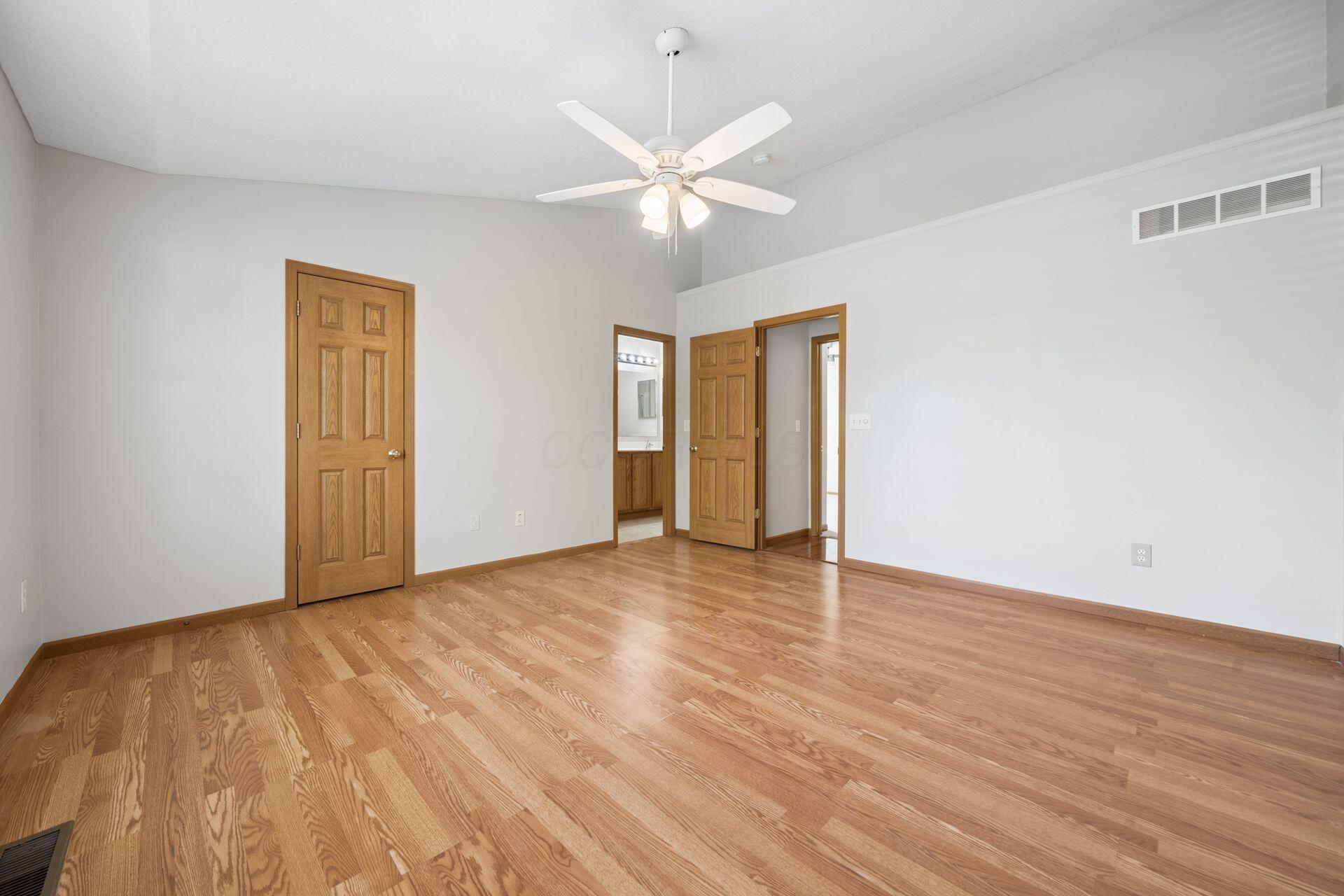 Empty bedroom with hardwood floors, a ceiling fan, and an open doorway to the bathroom.